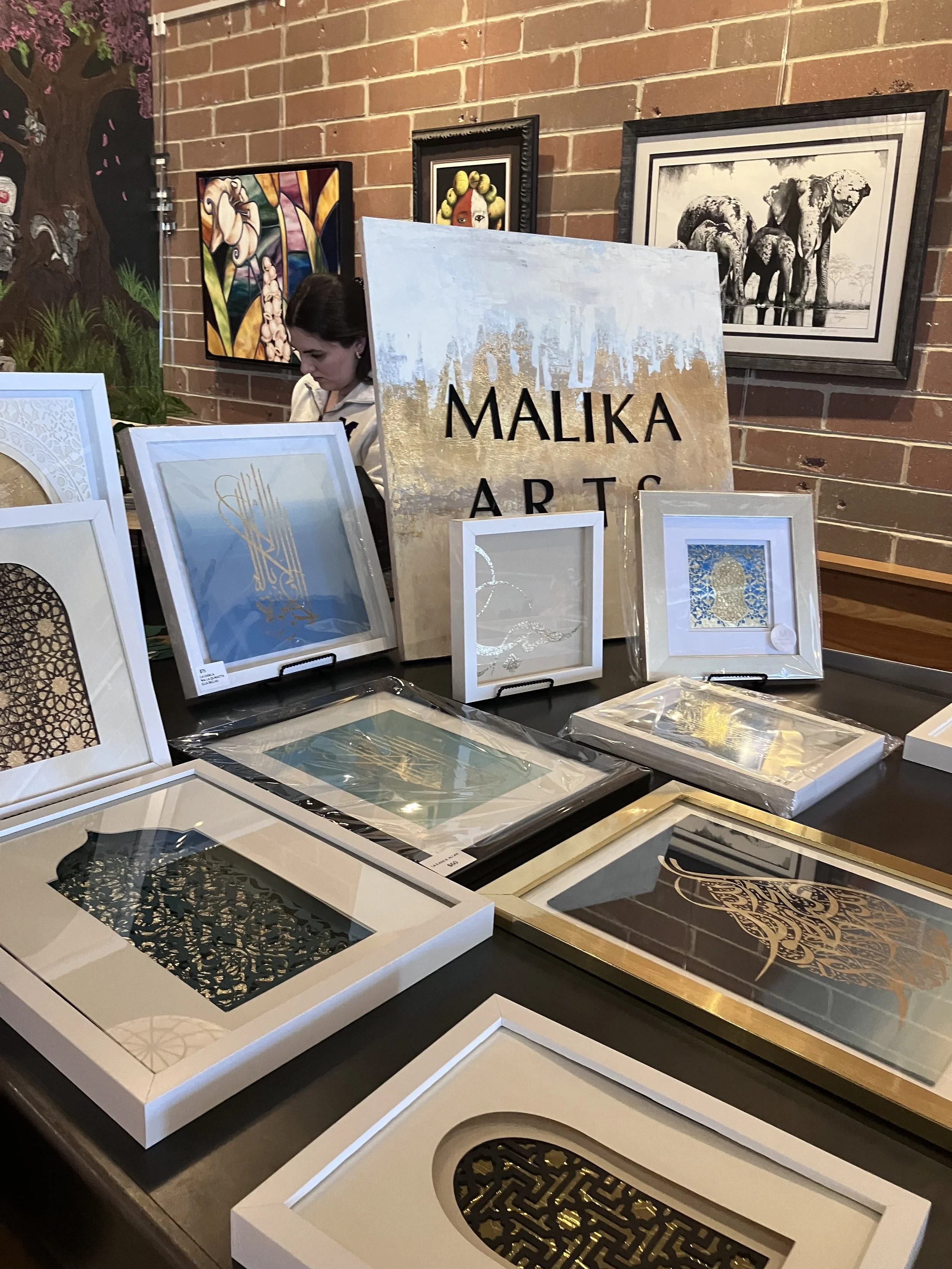 Artworks displayed on tables at an art exhibition with paintings and framed pieces, a woman browsing, and a brick wall in the background.