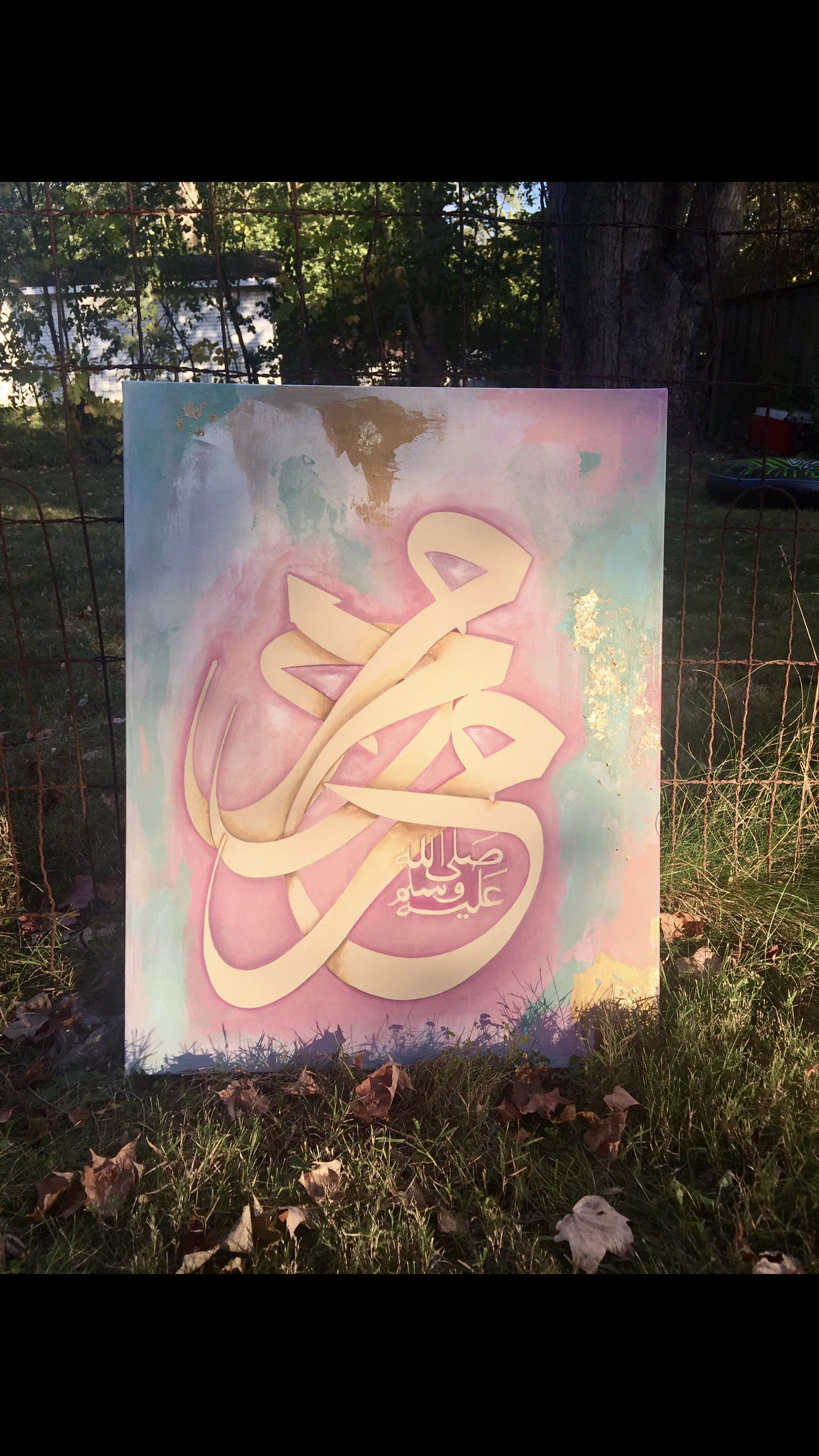 A colorful painting of Arabic calligraphy with a pink, teal, and gold background, displayed outdoors against a wire fence with a tree and grass in the background.