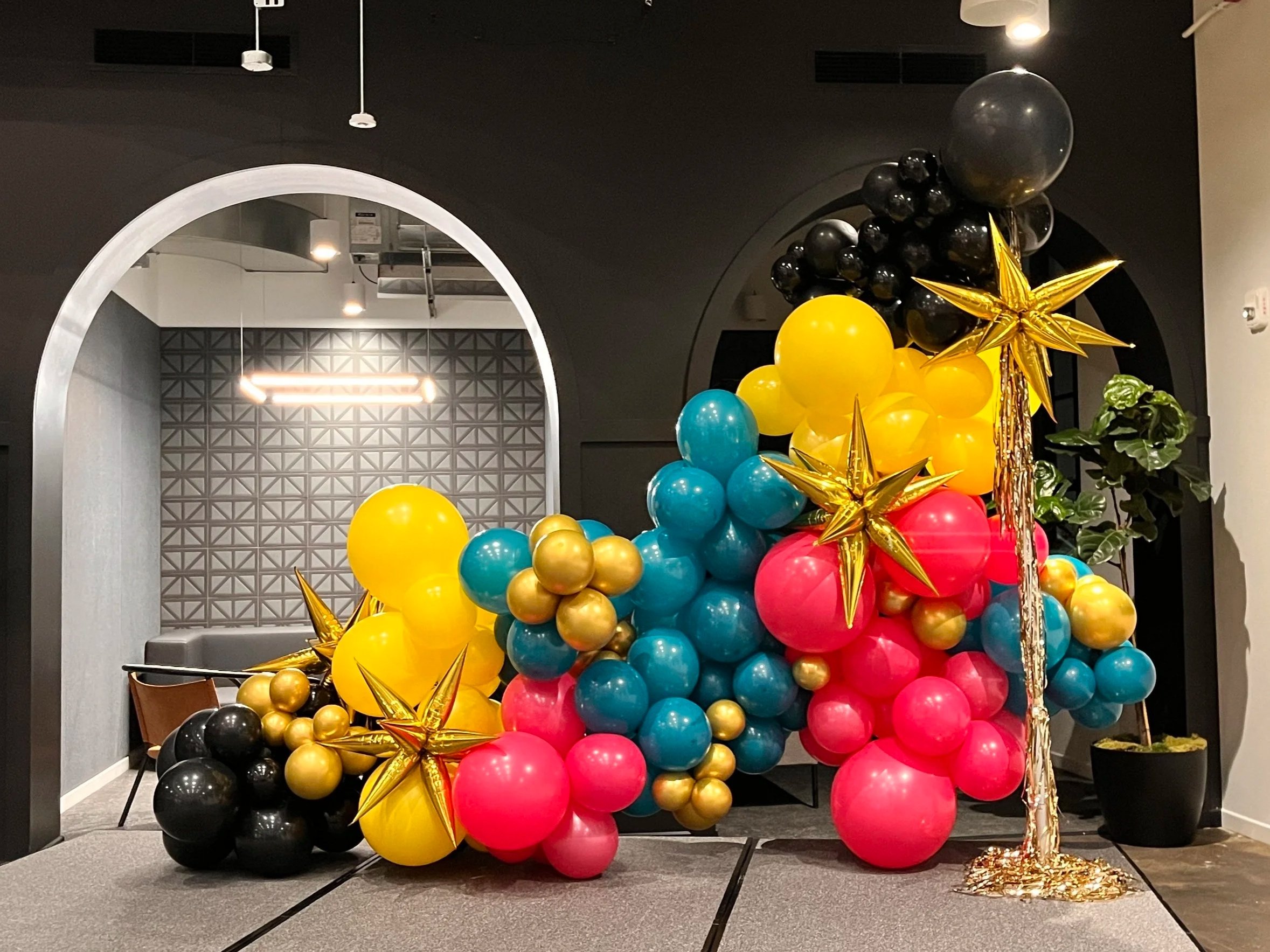 Colorful balloon arrangement with black, blue, yellow, pink, and gold balloons, some shaped as stars, in a modern interior with arched doorway and potted plant.