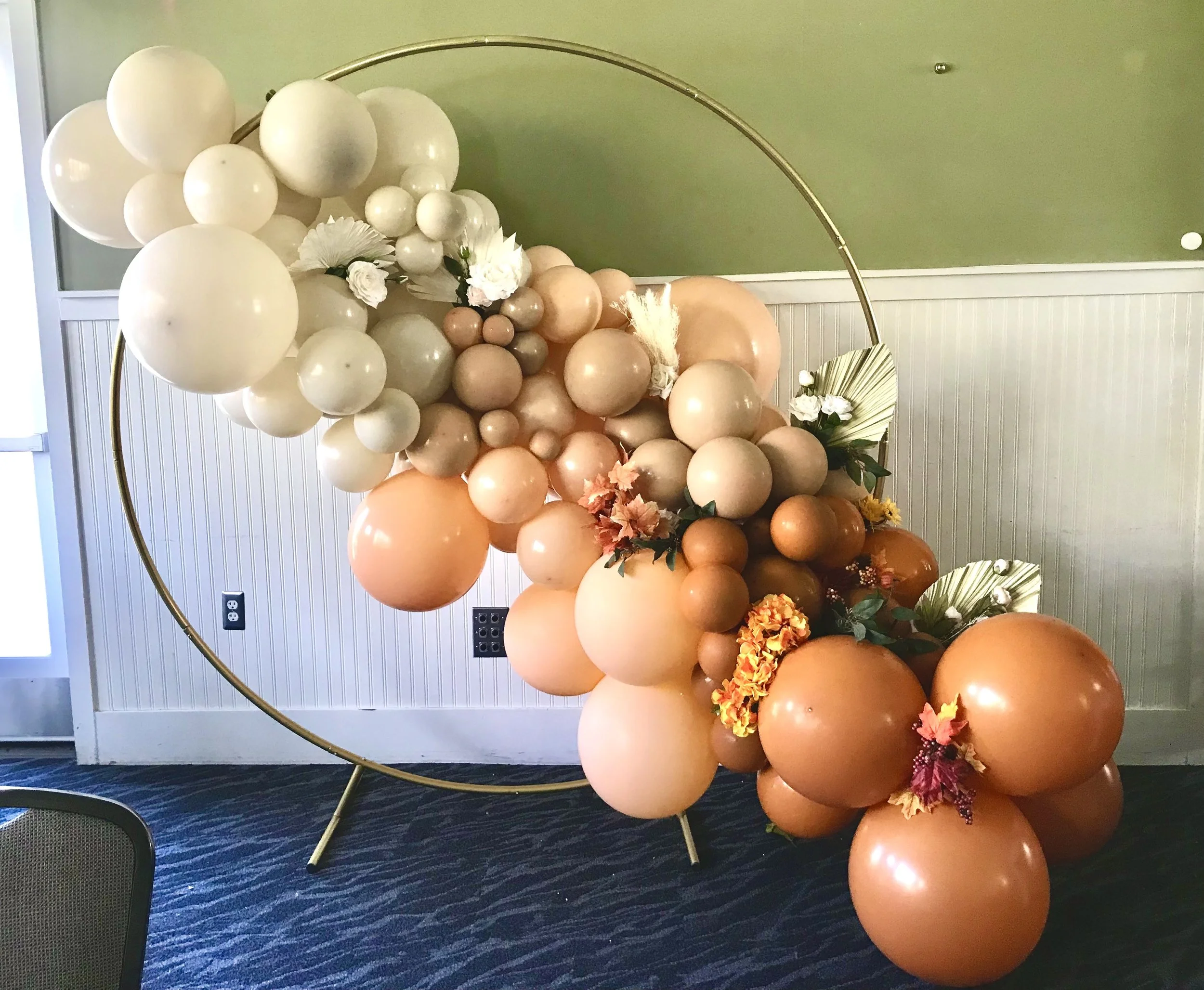 Decorative balloon arrangement with white, beige, tan, and orange balloons, accented with flowers, leaves, and a circular gold frame.