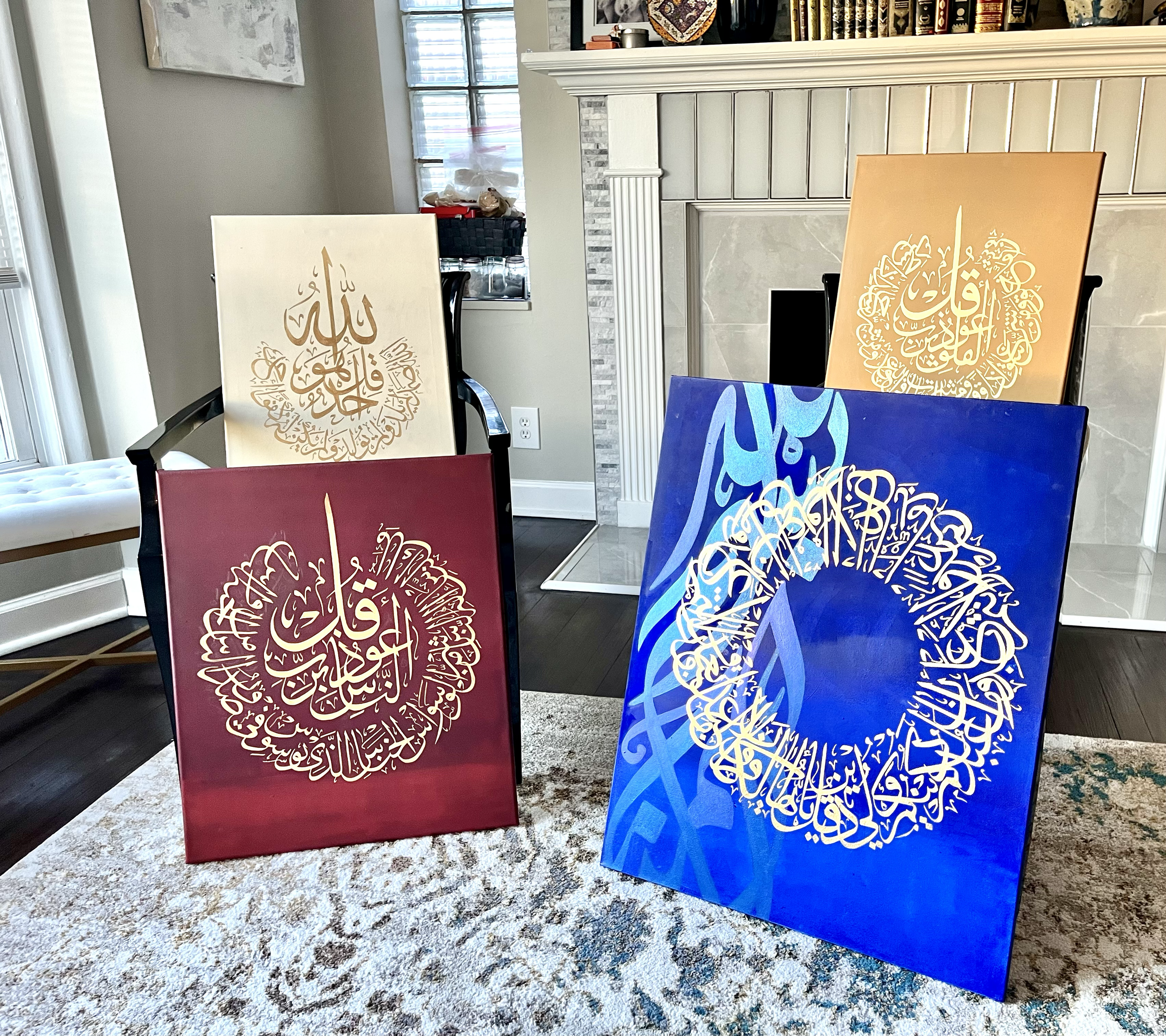 Four abstract paintings with intricate Arabic calligraphy in gold, blue, beige, and burgundy colors, displayed on stands in a living room with a fireplace and a window in the background.