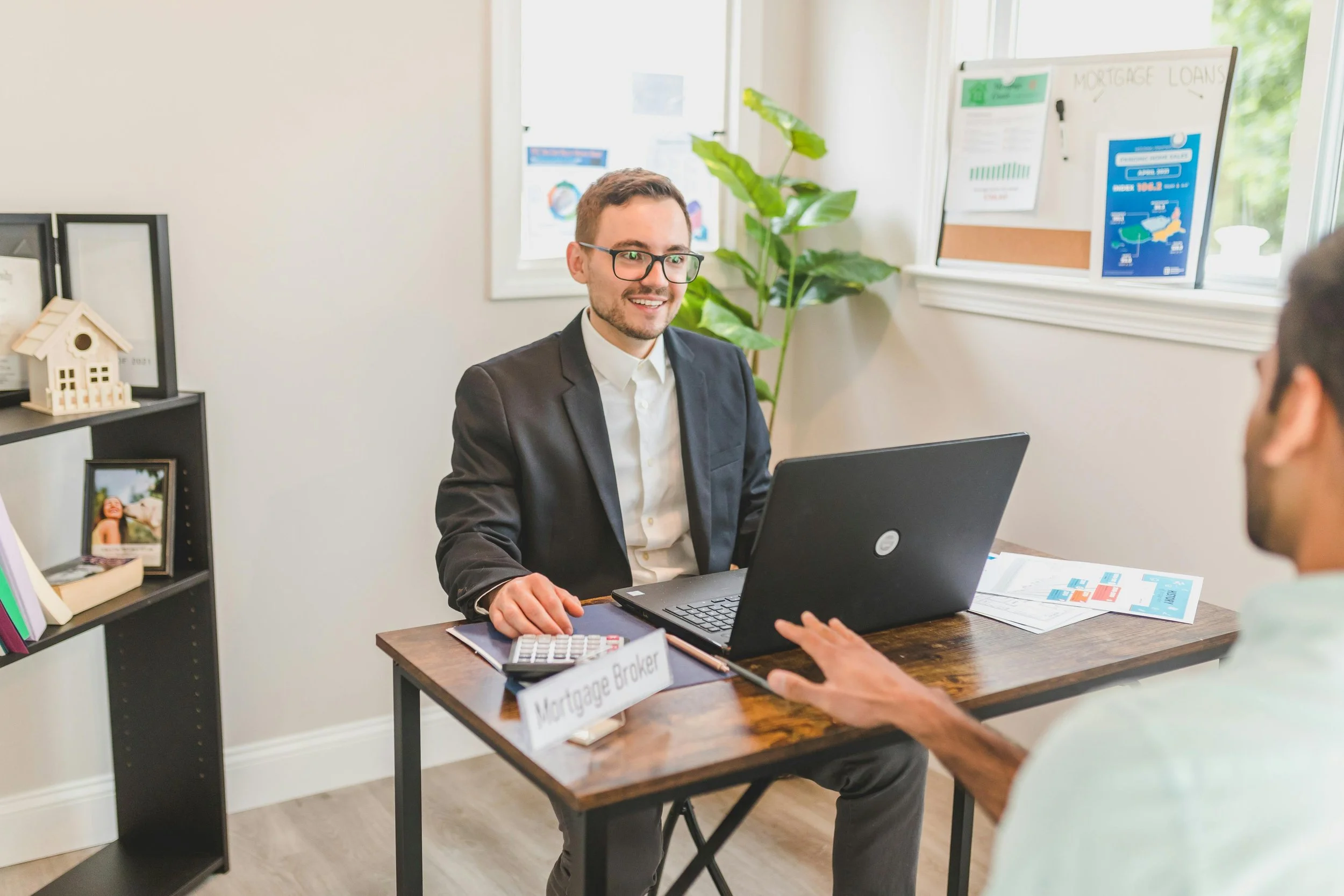 A man in a suit sitting at a desk in an office, talking to another person, with a sign that says 'Mortgage Broker' on the desk.