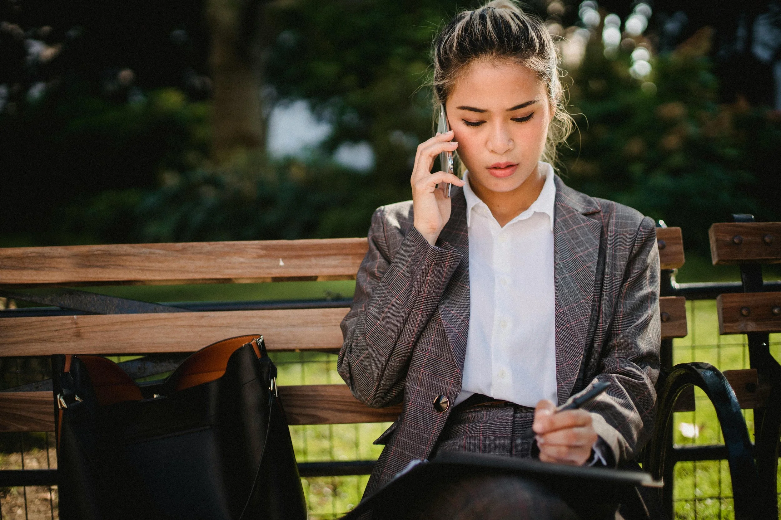 A young woman in business attire sitting on a park bench, talking on her cell phone and writing in a notebook.