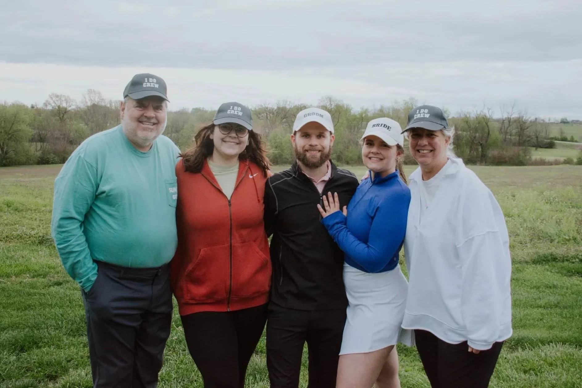 Group of five people outdoors on a cloudy day, celebrating a wedding, with the bride wearing a blue jacket and white skirt, and the groom in a black jacket and pink shirt, surrounded by family members wearing hats with wedding labels.