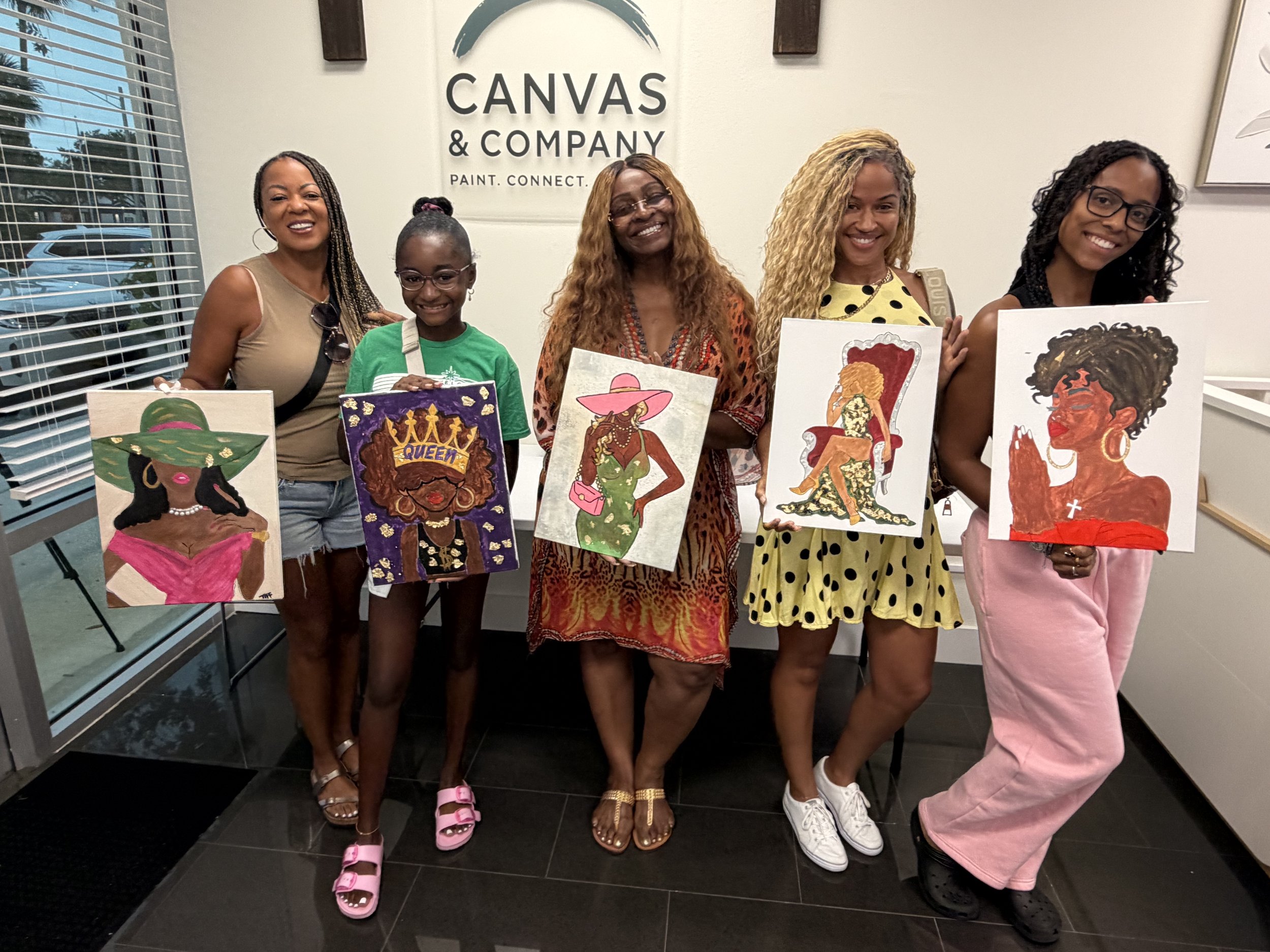A group of five women and one girl holding colorful paintings of women, standing in front of a wall with the logo and name of Canvas & Company. The women are smiling and dressed casually, showing a variety of hairstyles and clothing styles.