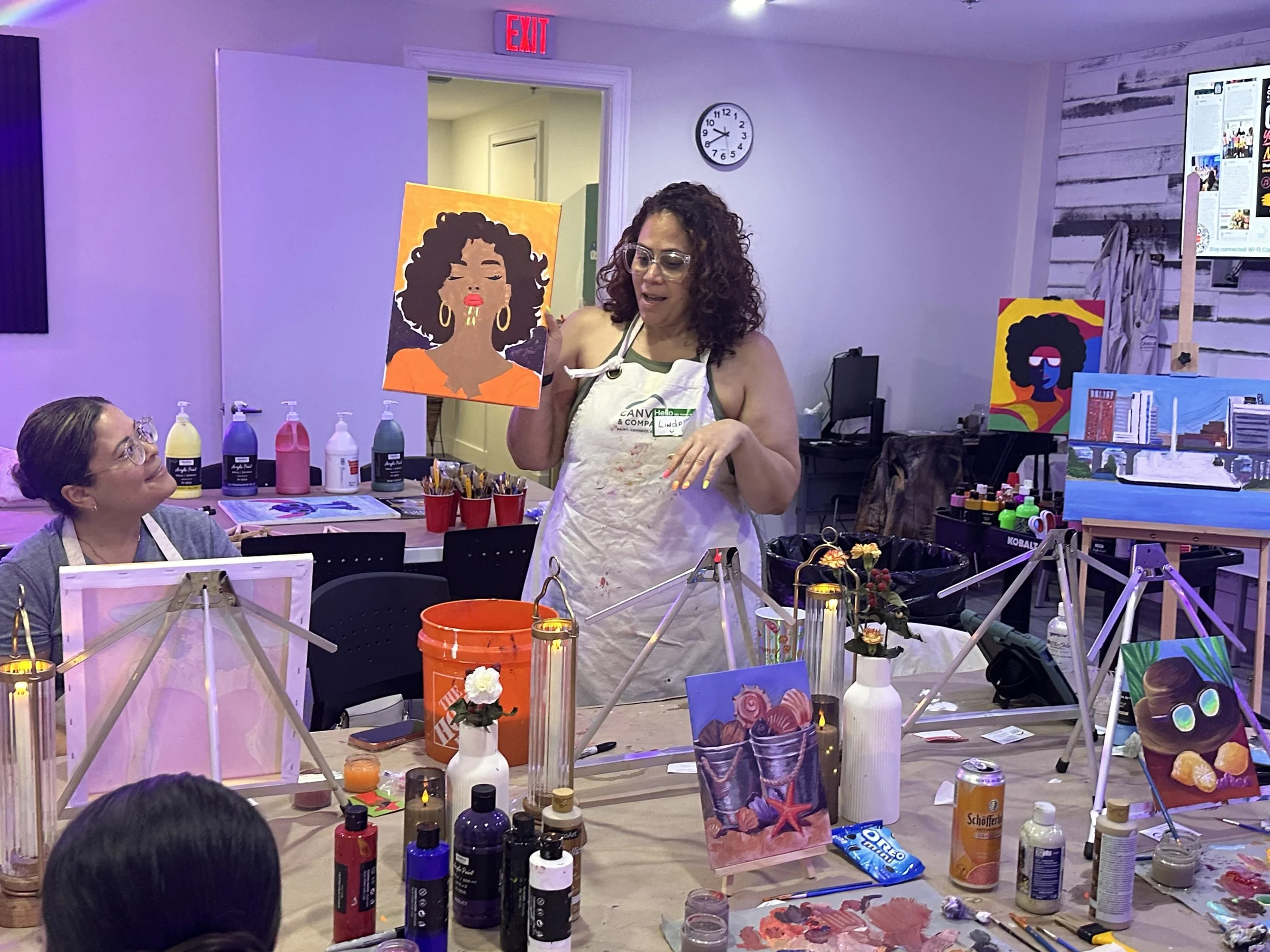 A woman in a painting class holding up a finished portrait of a woman with curly hair, gold hoop earrings, and red lipstick.