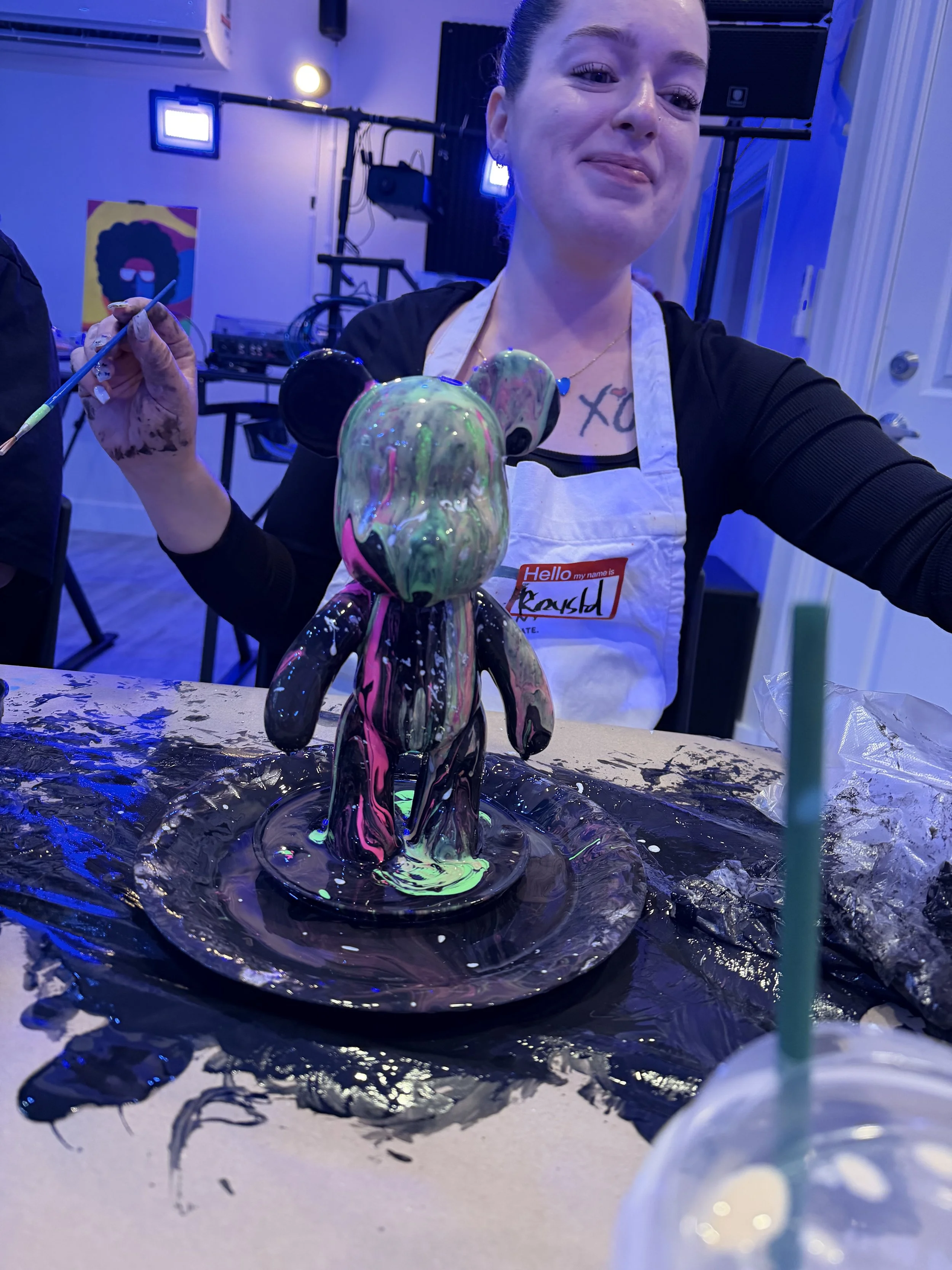 A woman in a white apron painting a multicolored, abstract teddy bear sculpture at an art event with blue lighting.