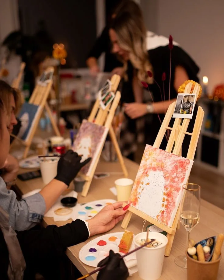 People participating in a painting activity at a wine and paint event, with small canvases, paint palettes, and brushes on tables.