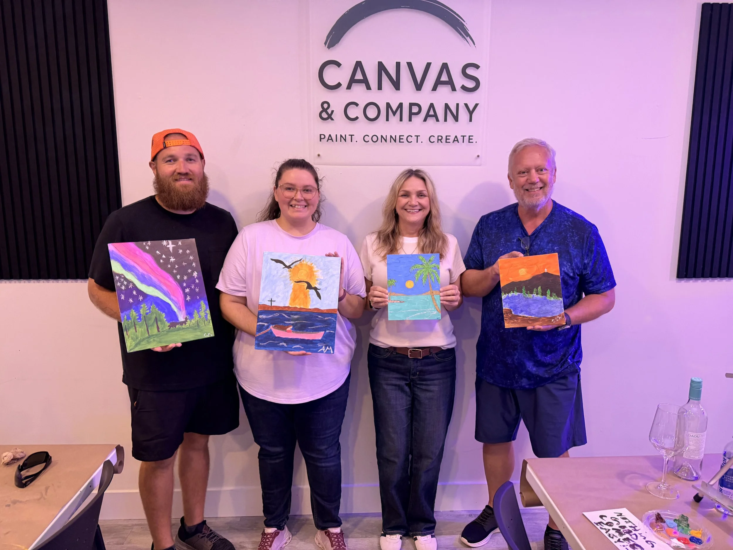 Four people standing indoors against a wall with a sign that reads, "CANVAS & COMPANY PAINT. CONNECT. CREATE." Each person is holding colorful paintings they created.