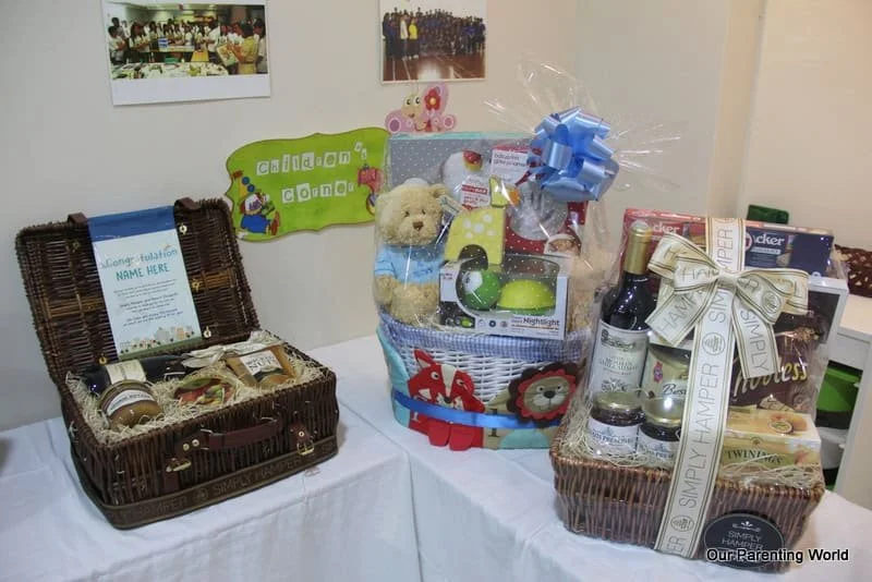 Table with three baskets of gifts, including stuffed animals, bottles, and jars, against a wall with children's photos and a colorful sign reading "Child's Note."