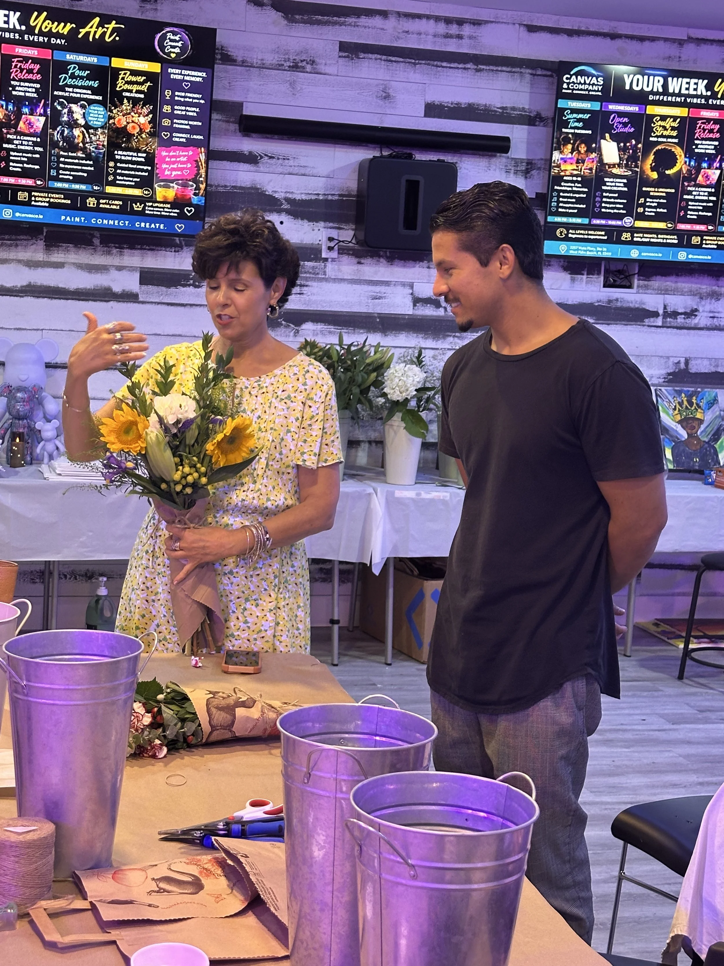 A woman in a yellow floral dress holding a bouquet of sunflowers and white flowers, standing next to a man in a black t-shirt and plaid pants, in a room decorated with flowers and arts and crafts supplies, with large screens displaying event schedules in the background.