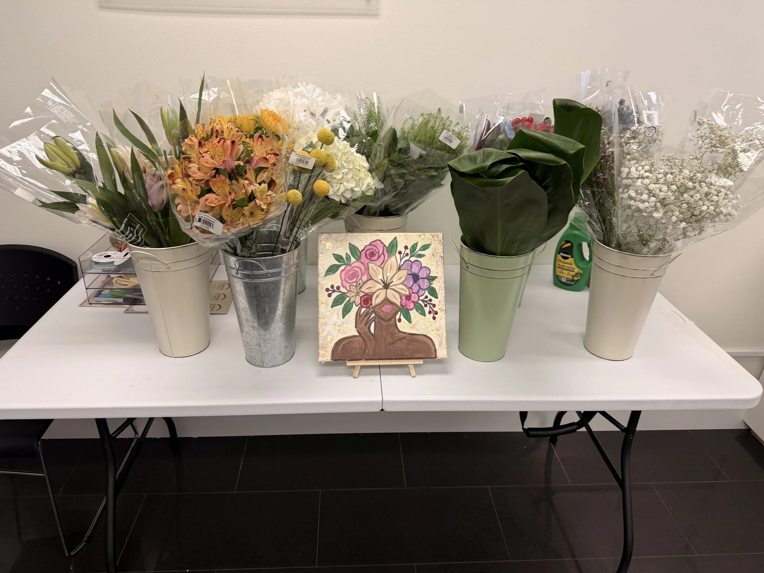 Several bouquets of flowers placed in white and metal buckets on a table, with a small colorful floral painting in front; a bottle of organic plant food and a pair of scissors are also on the table.