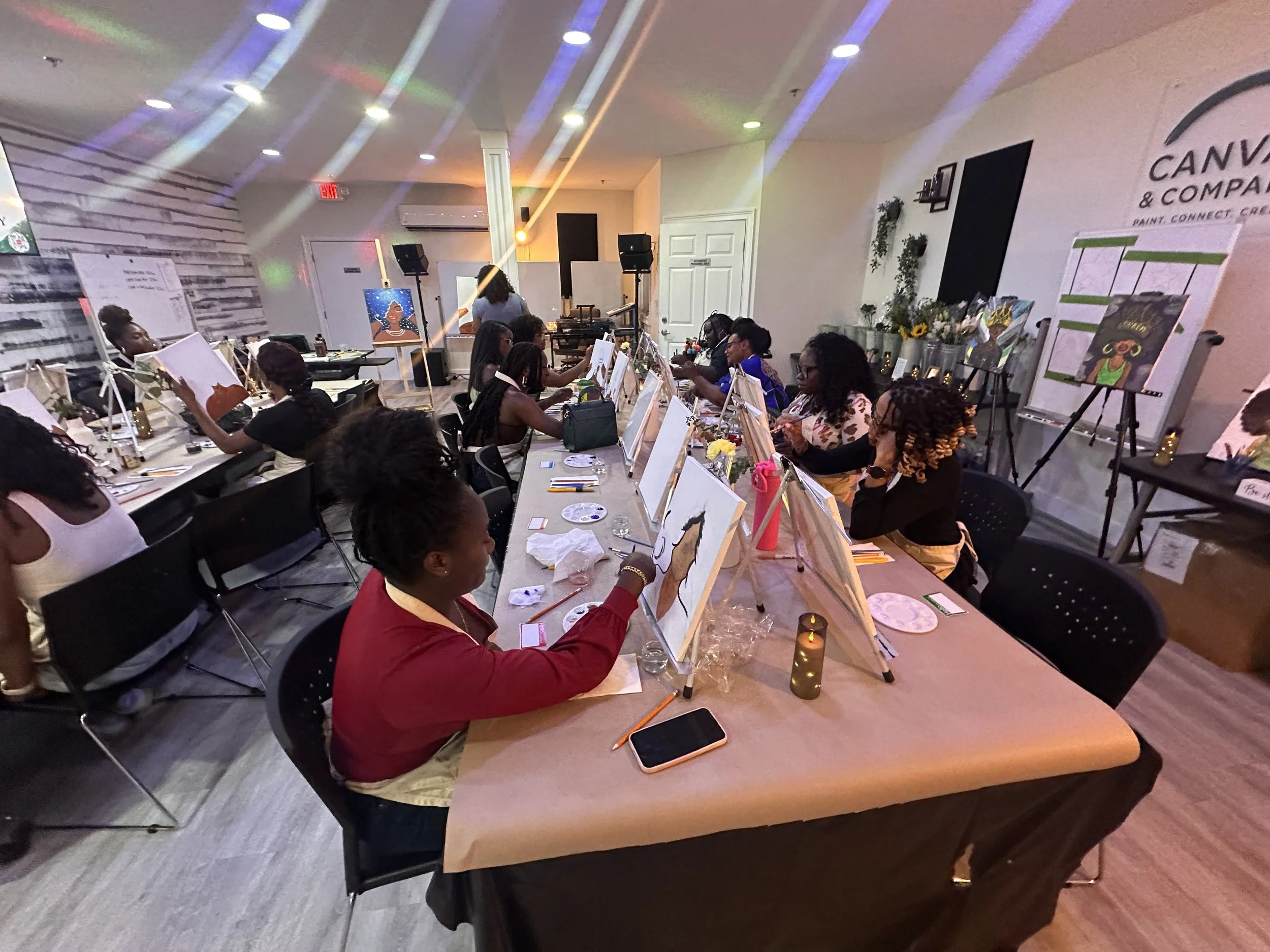 A group of people participating in a painting class, sitting bubbleside at a long table with art supplies and canvases, with colorful lights and artwork on display in the background.