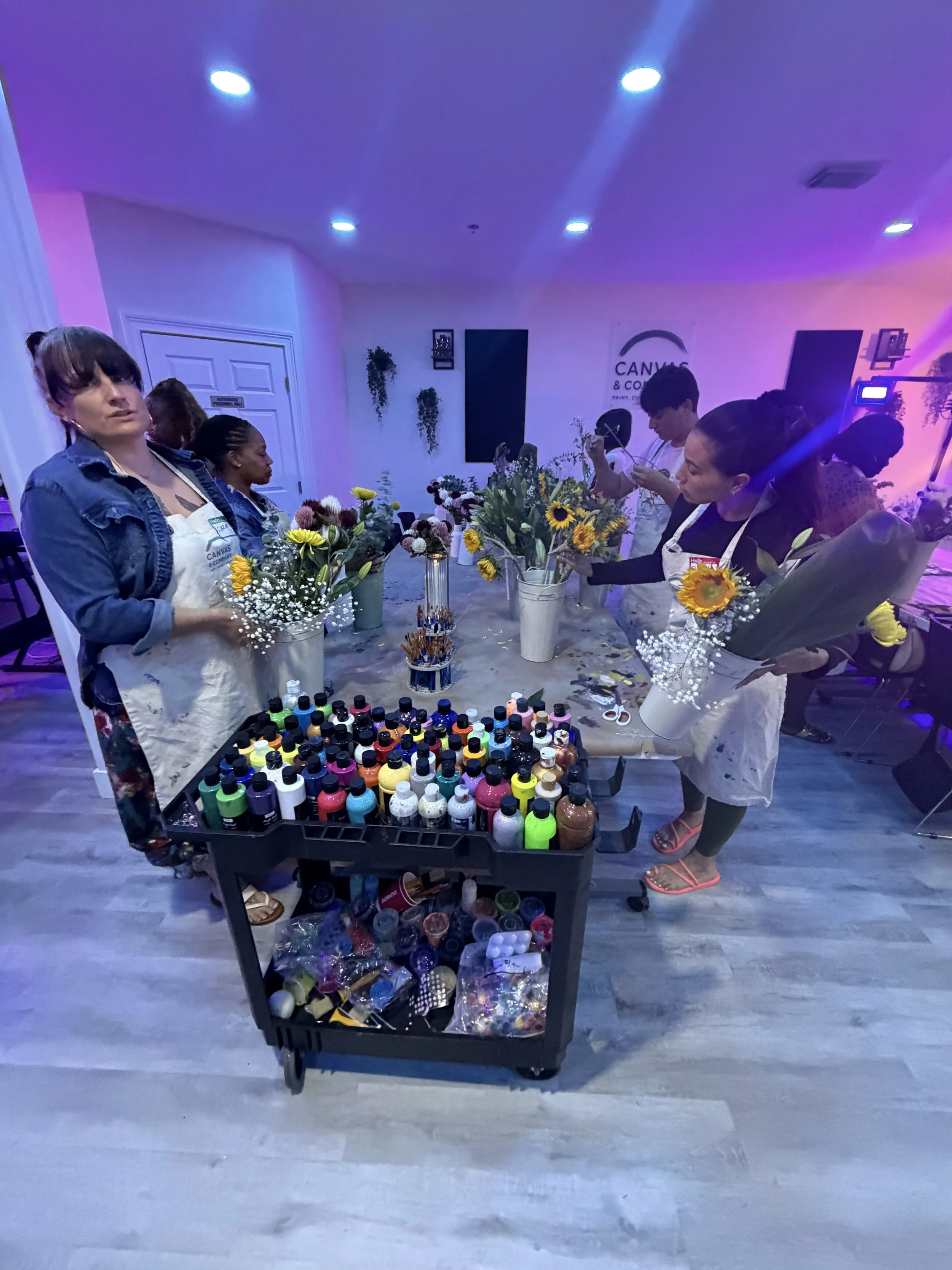 People preparing floral arrangements with colorful flowers and paint bottles at an event in a studio.