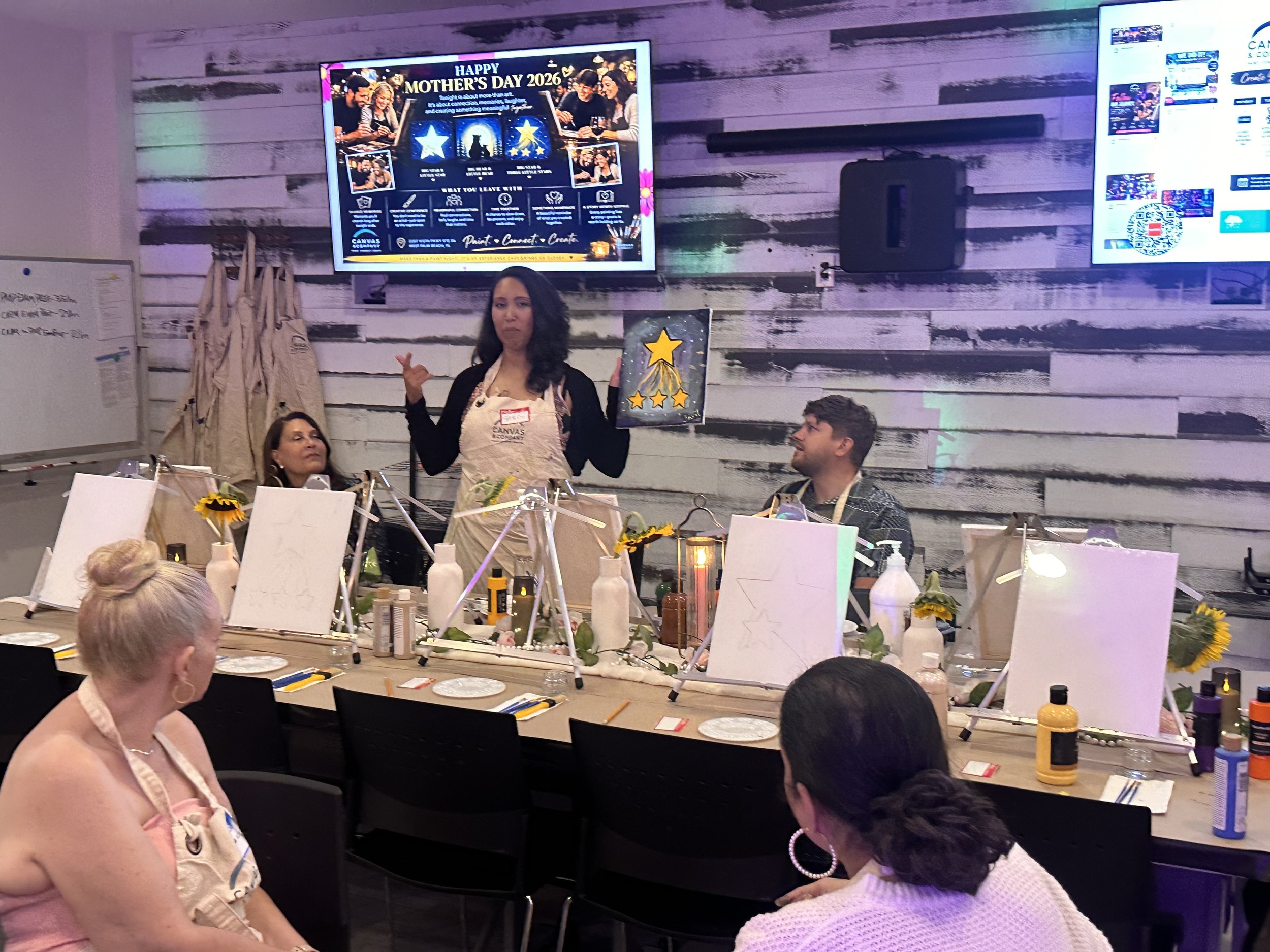 A group of women and a man participating in a painting activity at a Mother's Day event, with a man standing in front of the table pointing to the camera, in a room with wooden wall paneling and two large screens displaying Mother's Day graphics.