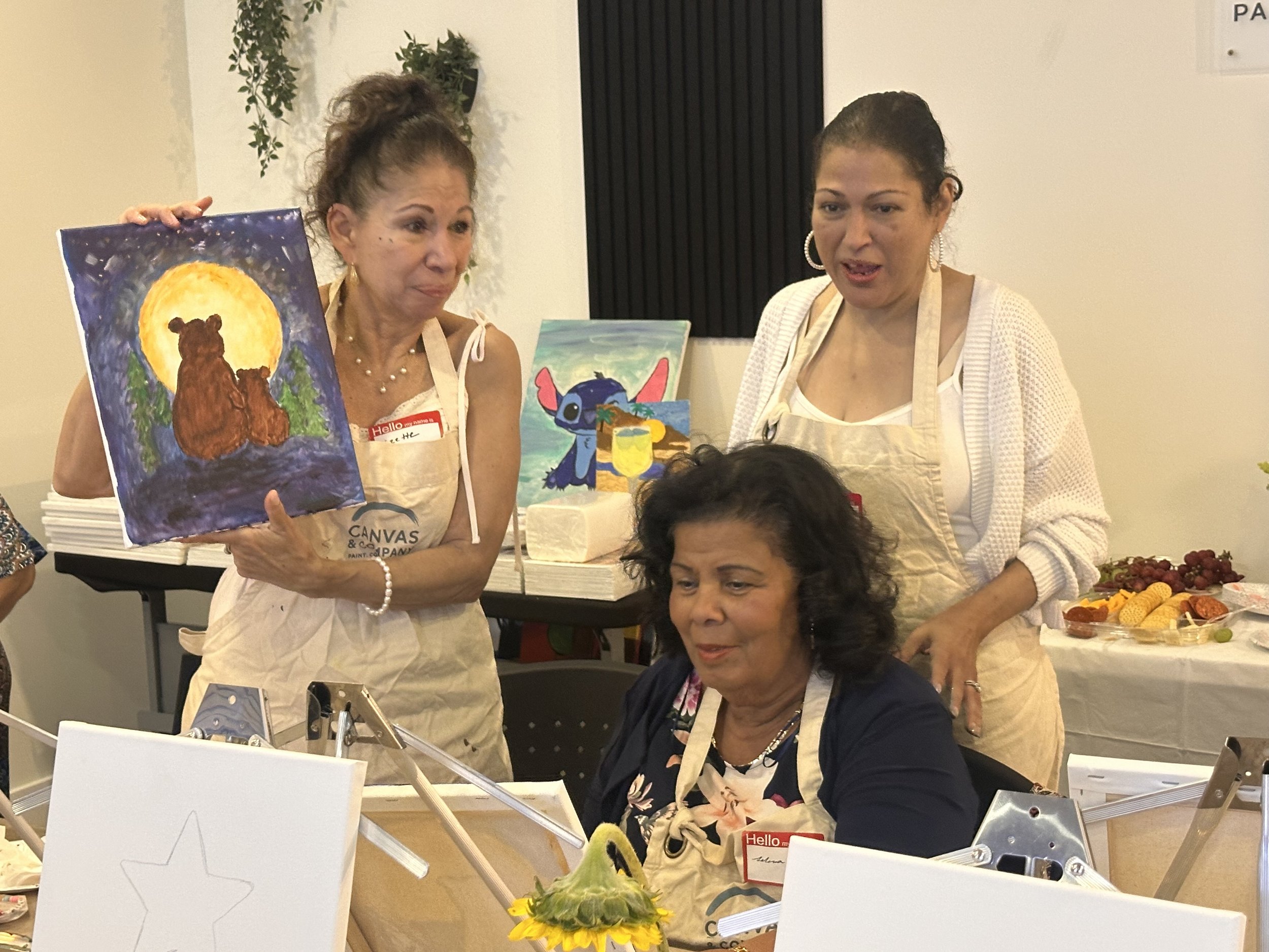 Two women at an art event, one holding a painting of two bears sitting near a large yellow moon, with a second woman in a white sweater and large earrings looking on.