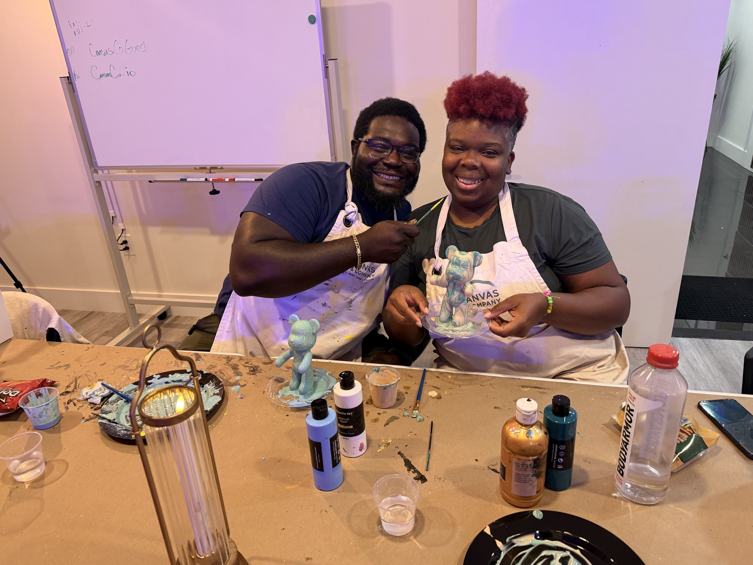 Two people sitting at a table during a pottery painting activity, holding a painted ceramic figure. The table is cluttered with various paint bottles and supplies, and a whiteboard is visible in the background.