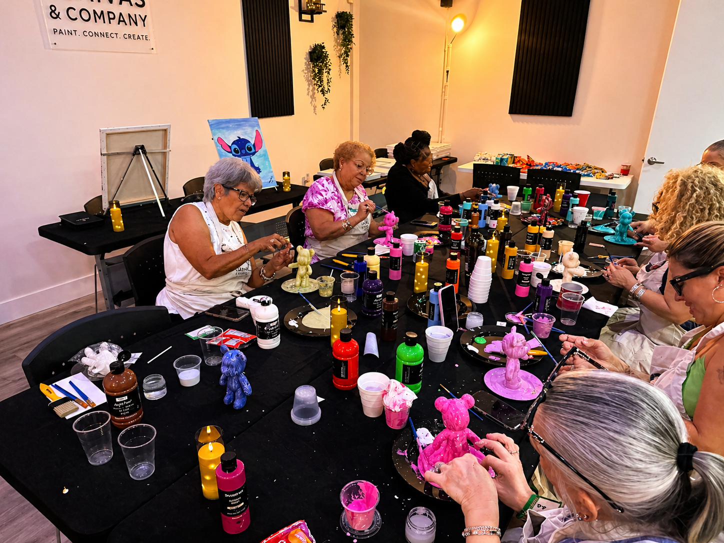 Women participating in a sip and paint event, painting colorful bear-shaped figurines, with art supplies and painted figures on the table in a well-lit room.
