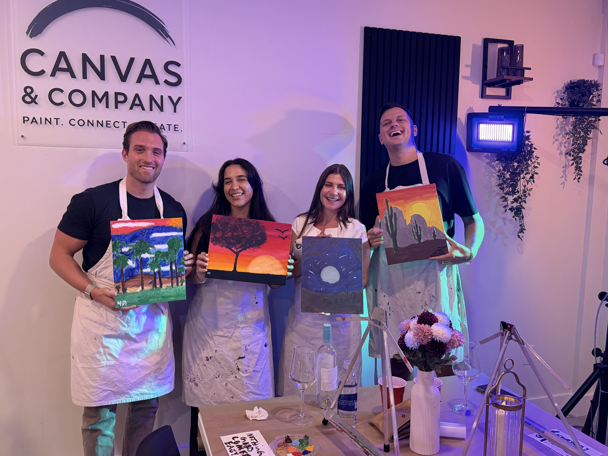 Four people smiling and holding colorful paintings at an art event in a room with a sign reading 'CANVAS & COMPANY' on the wall.