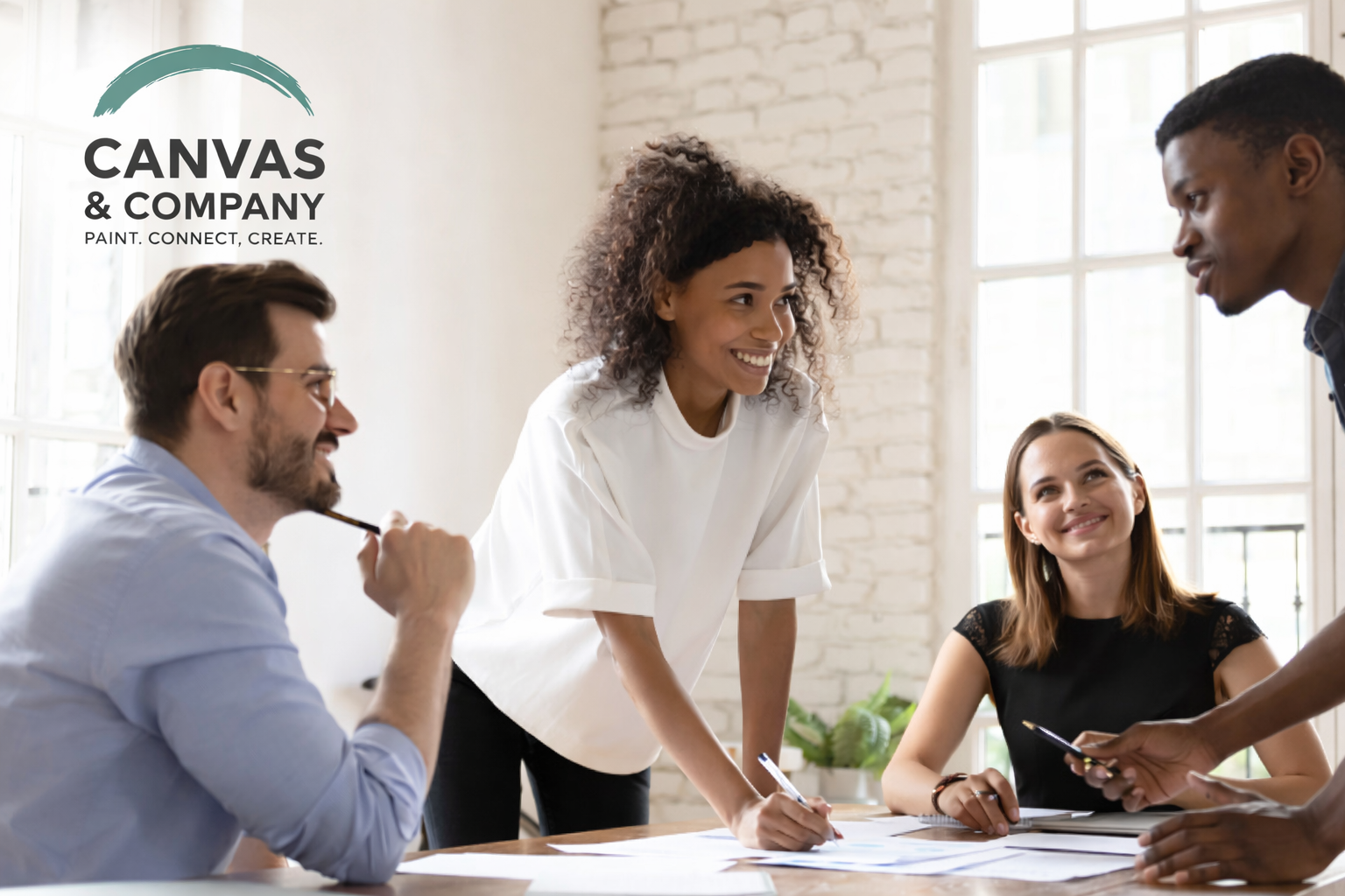 Group of diverse young professionals collaborating in a bright modern office with large windows, discussing and working on documents.