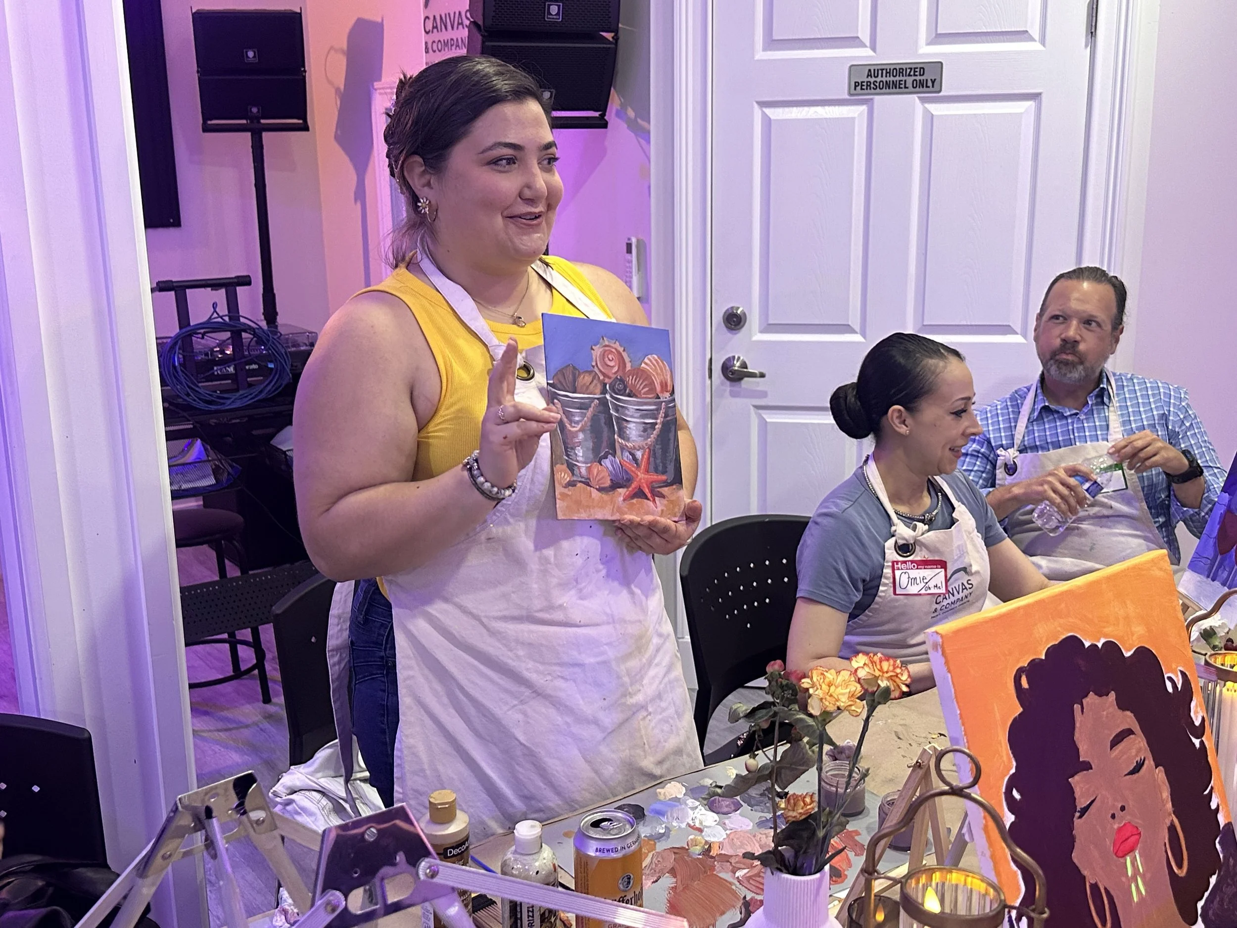 A woman in a yellow top and earrings holding up a colorful painting at an art event. Two people sitting at a table are painting and working with art supplies, with a pink and purple ambient lighting in the background and a sign on the door that reads 'AUTHORIZED PERSONNEL ONLY'.