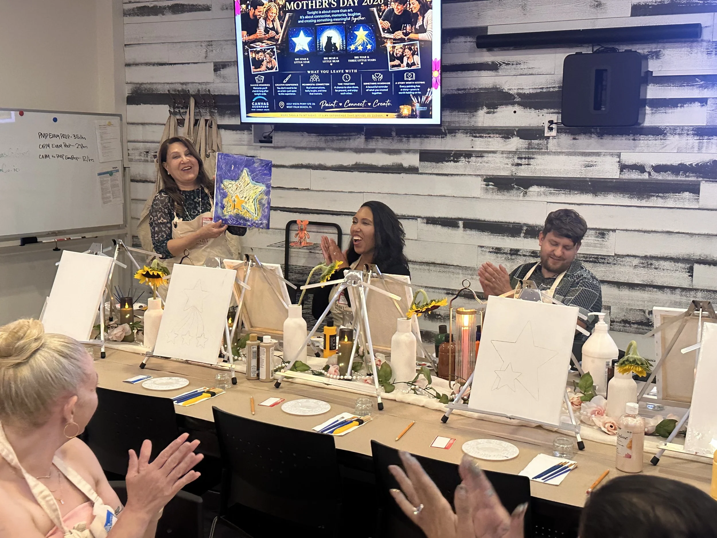 Women and men celebrating Mother's Day in a painting class. A woman stands holding a painted canvas with a star design, smiling. Other participants are seated, clapping, and smiling at the event. The table is decorated with sunflowers, fairy lights, and art supplies. A digital display on the wall shows a Mother's Day event poster.