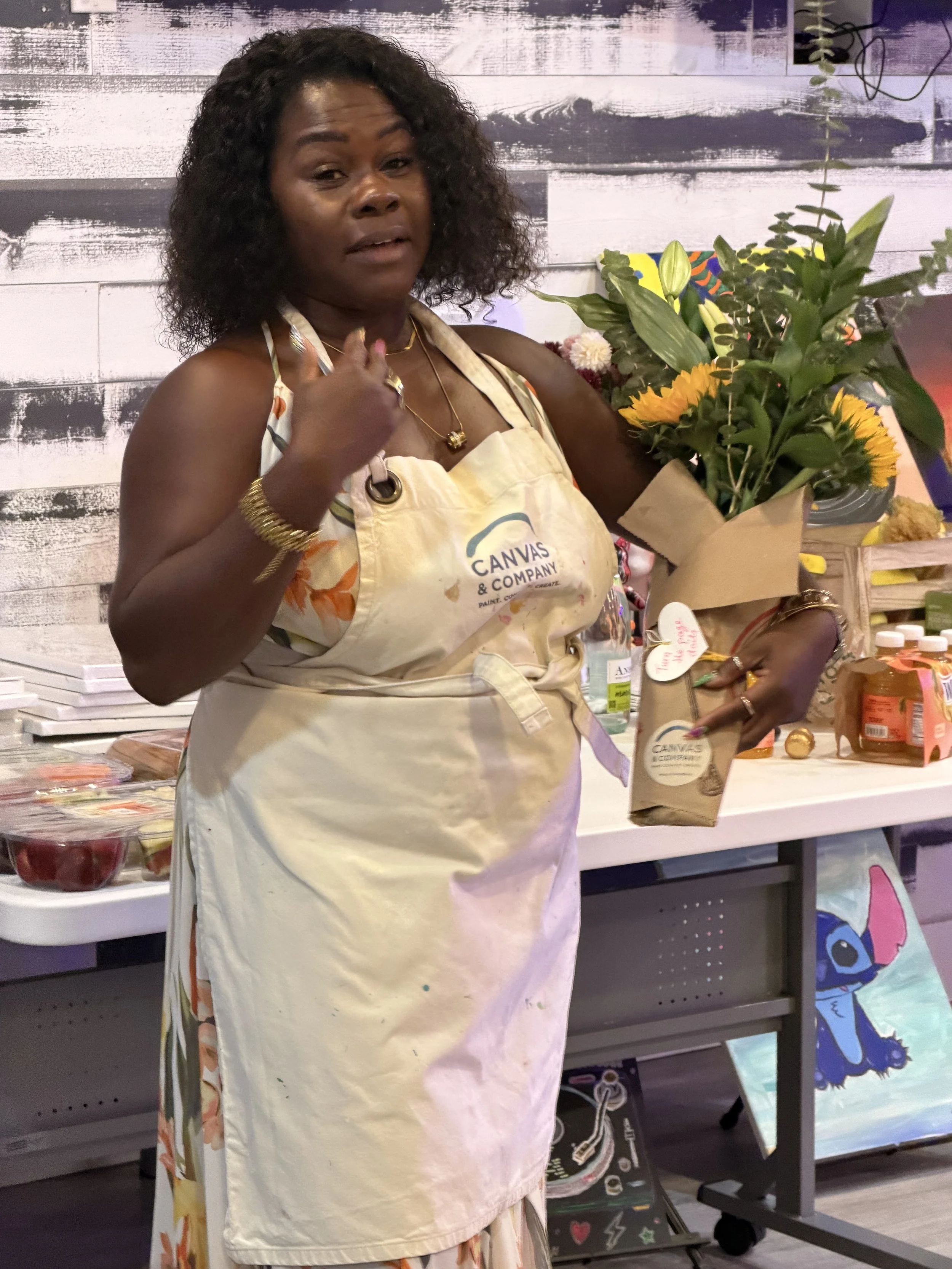 A woman wearing a beige apron with the logo 'Canvas & Company' holding a bouquet of flowers in brown paper. She is standing in front of a decorated wall and a table with art supplies and artwork.