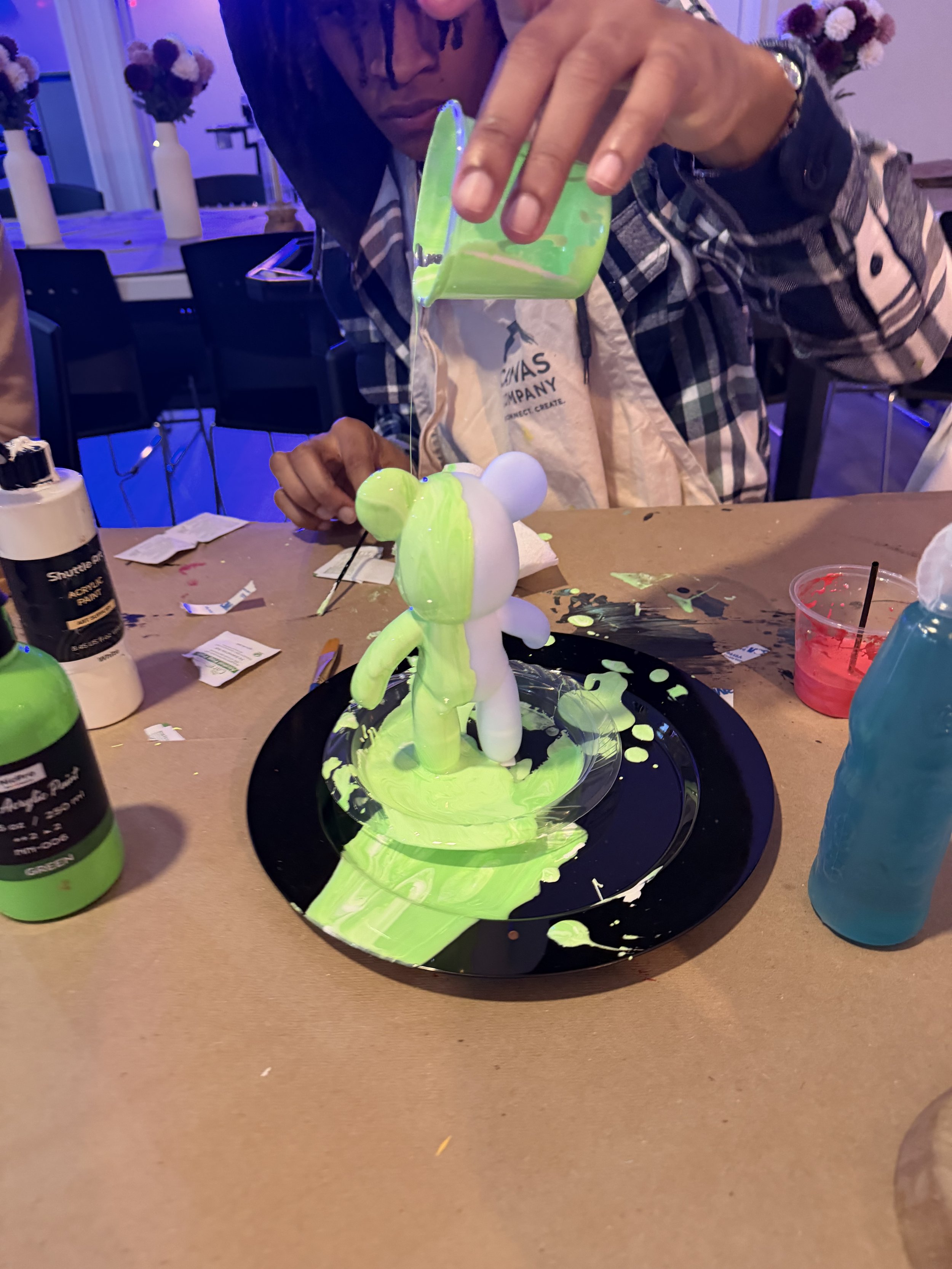 A person holding a green container pouring a bright green liquid over a white figurine on a black plate, with a table filled with art supplies and decorated with vases of flowers in the background.