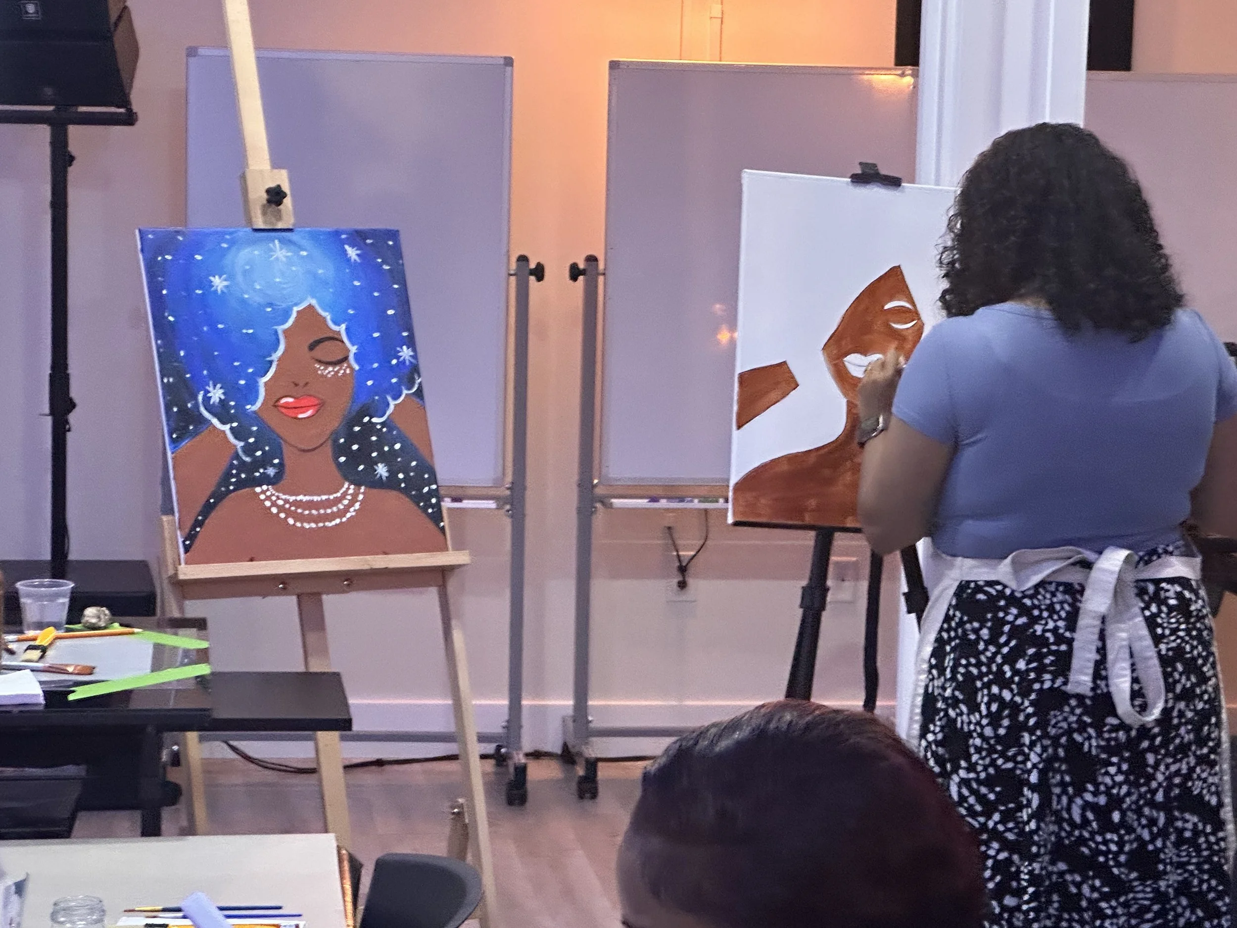 An artist is painting a portrait of a woman with blue curly hair, closed eyes, wearing pearl jewelry, on a canvas in a studio. Another artwork of a face with brown hair is visible on an easel to the side.