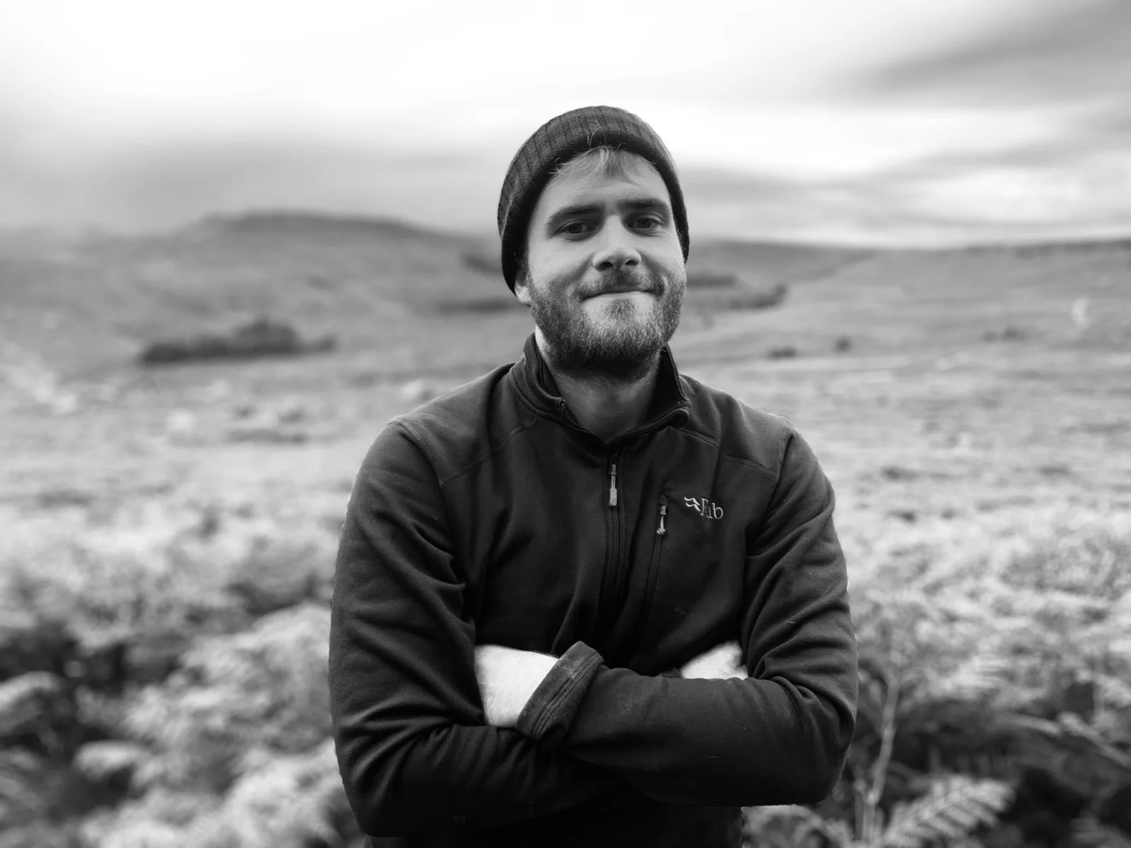 Black and white photo of a young man with a beard, wearing a beanie and a zipped jacket, standing with arms crossed outdoors in a hilly landscape with an overcast sky.