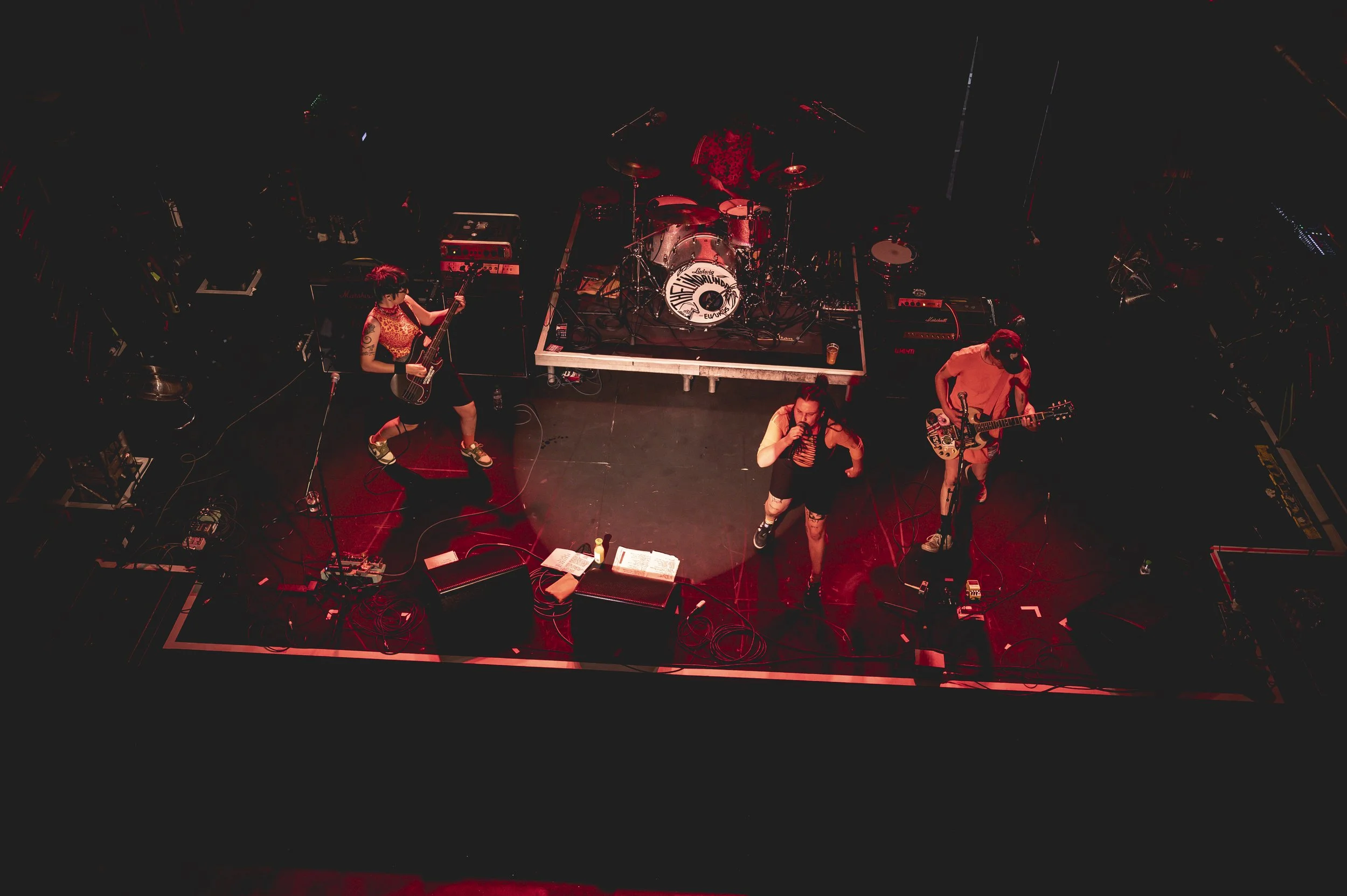 A band performing on a dimly lit stage with red lighting. Four members are visible: one playing bass, one singing into a microphone, and two playing guitars. A drum set is in the background. The stage has various equipment and cables.