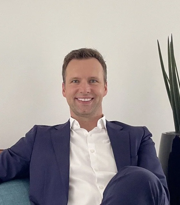 Ein lachender Mann in formeller Kleidung sitzt auf einem Sofa vor einem weißen Hintergrund, mit einer Pflanze im Hintergrund.