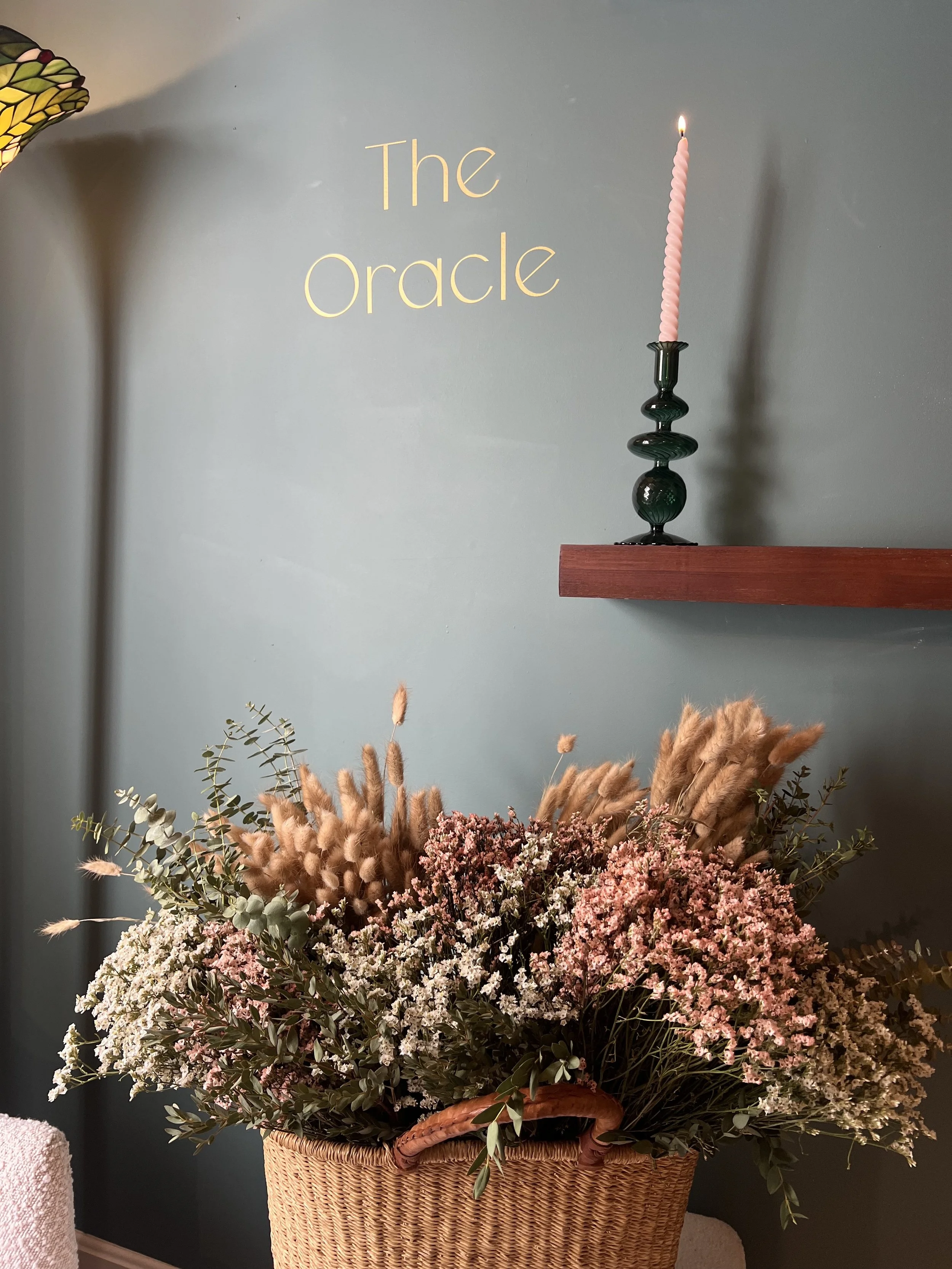 Decorative basket filled with pink and white flowers and greenery, located in front of a teal wall with the words 'The Oracle' written on it and a pink candle on a dark green candlestick on a wooden shelf
