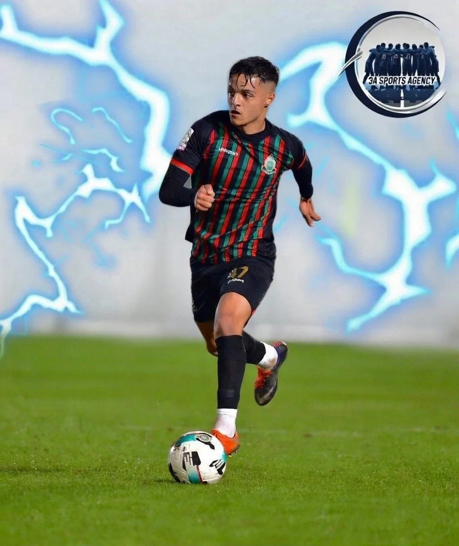 A young man playing soccer on a field at night, wearing a black uniform with red and green stripes, and kicking a soccer ball.