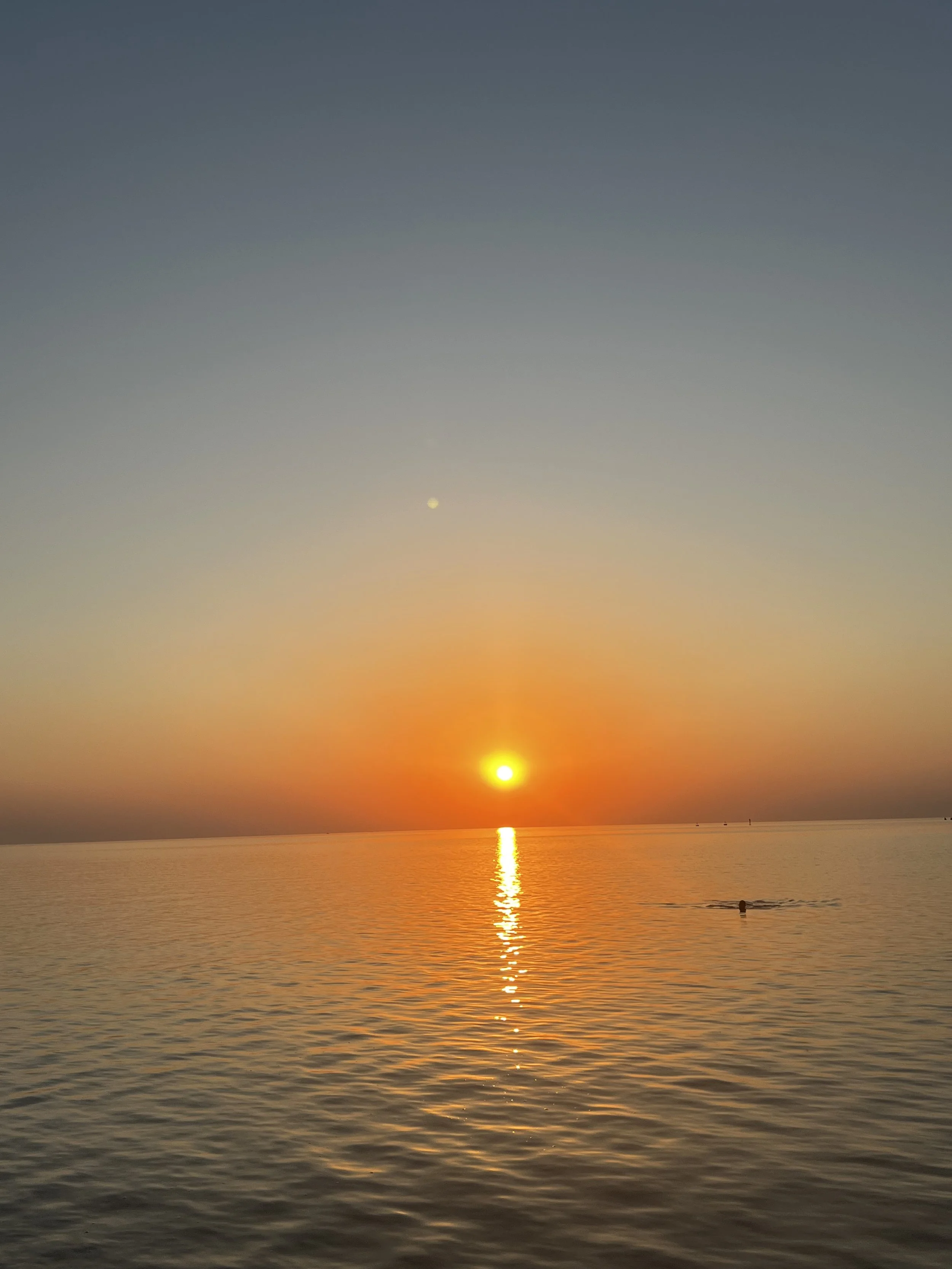 Sunrise over calm water with the sun reflected on the surface and a person swimming in the distance.