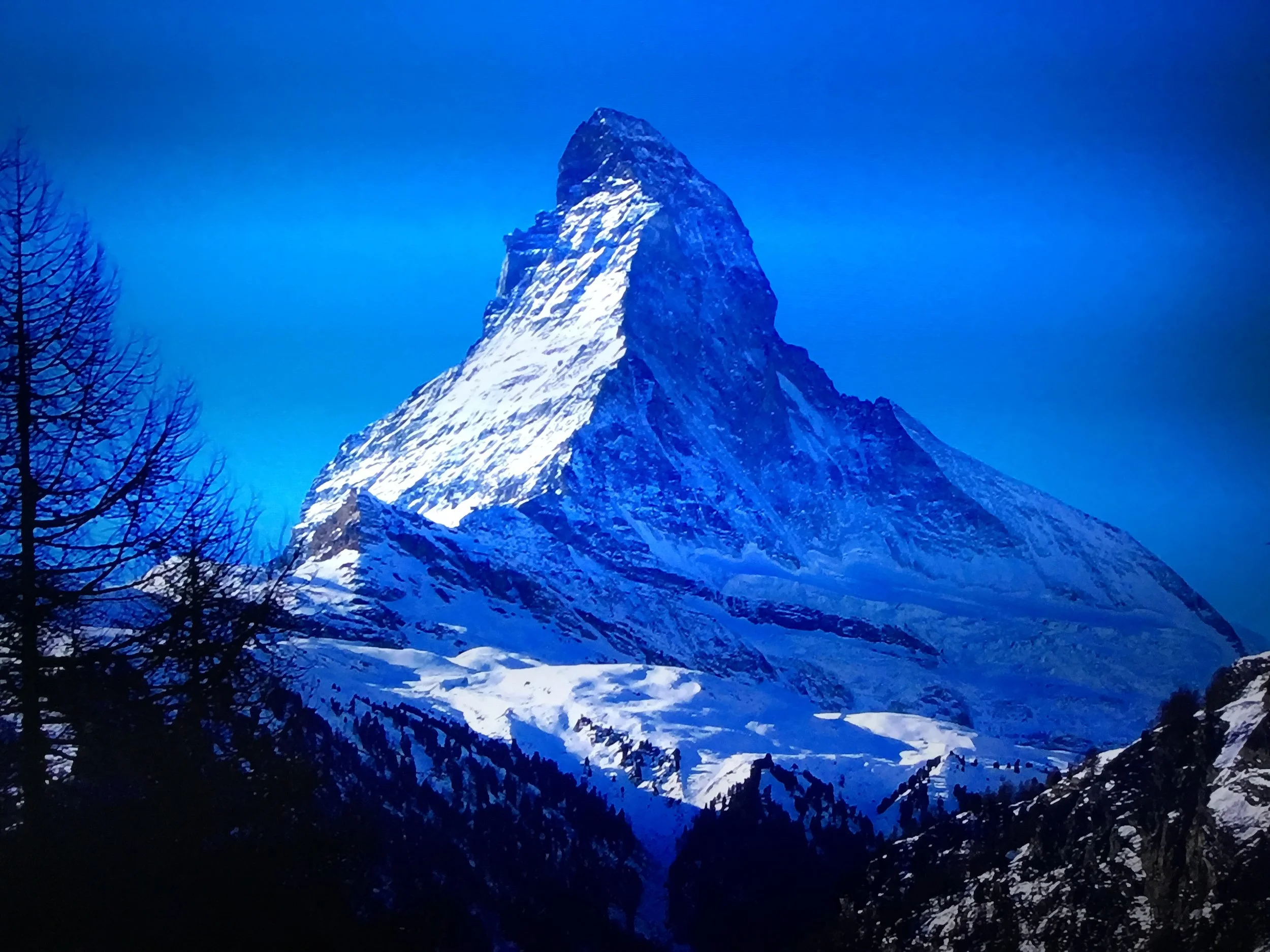Snow-covered mountain with sharp peak against a blue sky, with dark silhouette trees in the foreground.