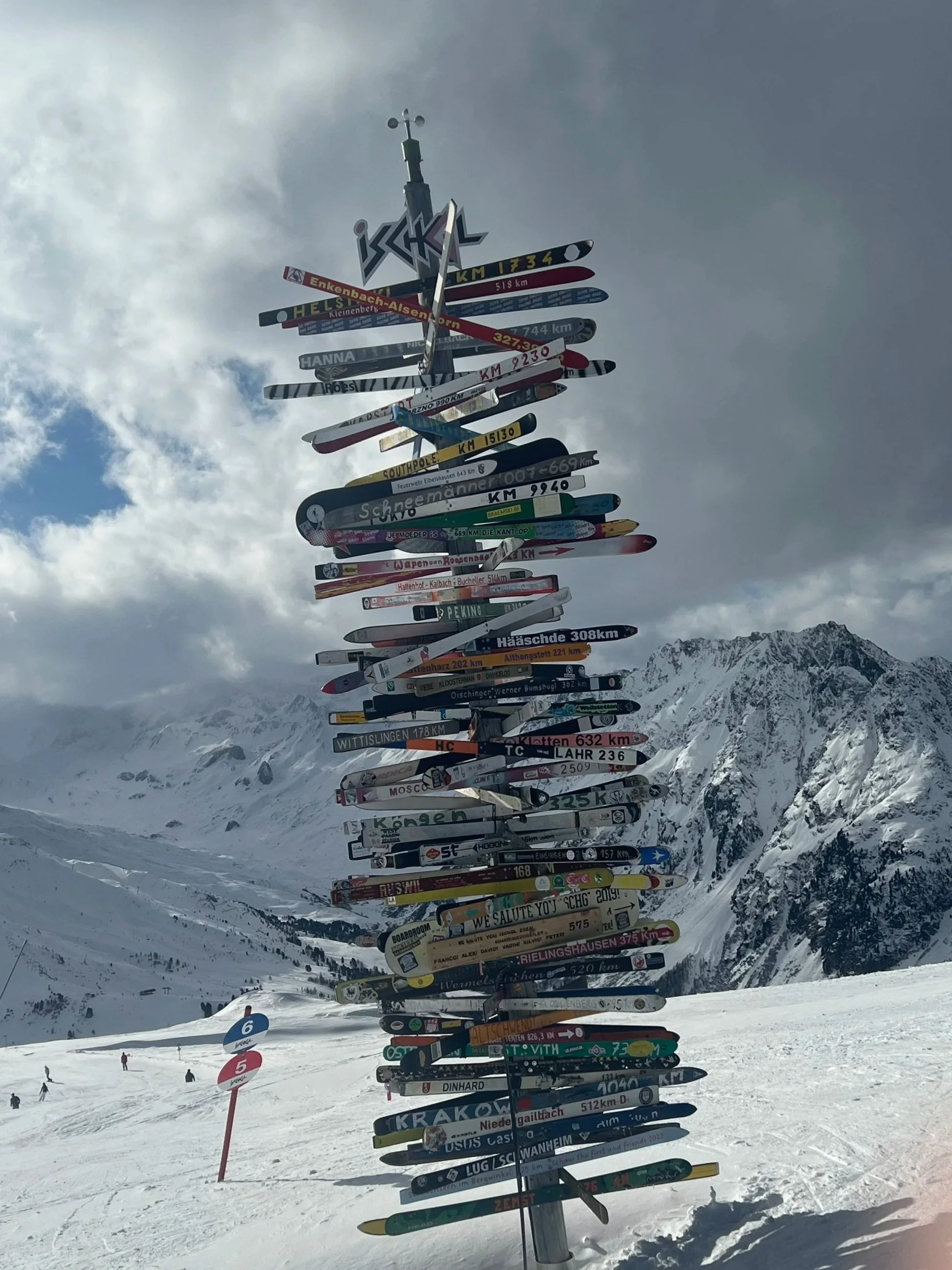 A ski pole with multiple colorful signs pointing in different directions, in a snowy mountain landscape.