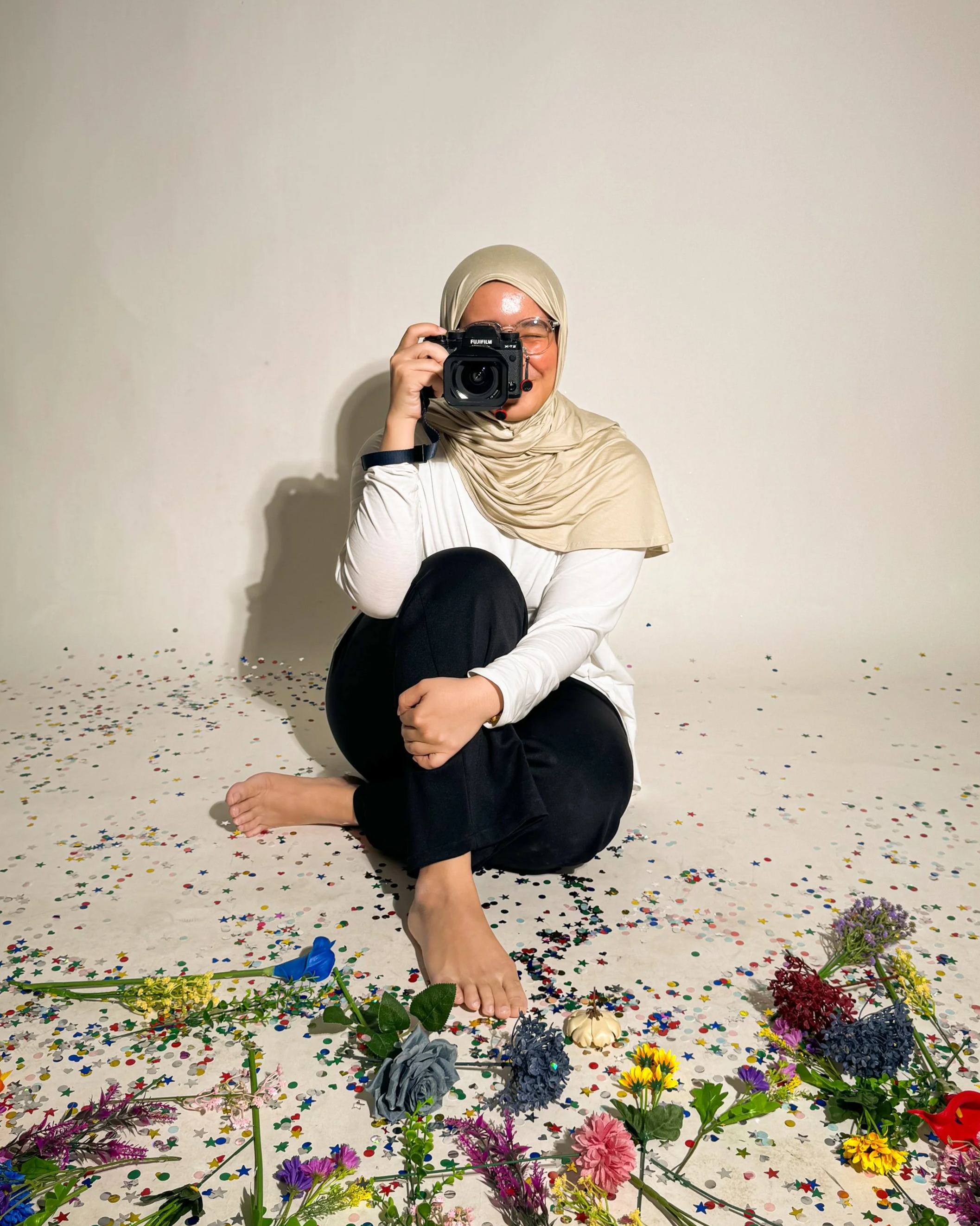A woman wearing glasses and a hijab is sitting on the floor, holding a camera to her eye, amidst scattered multicolored confetti and various flowers on the ground before a plain white background.