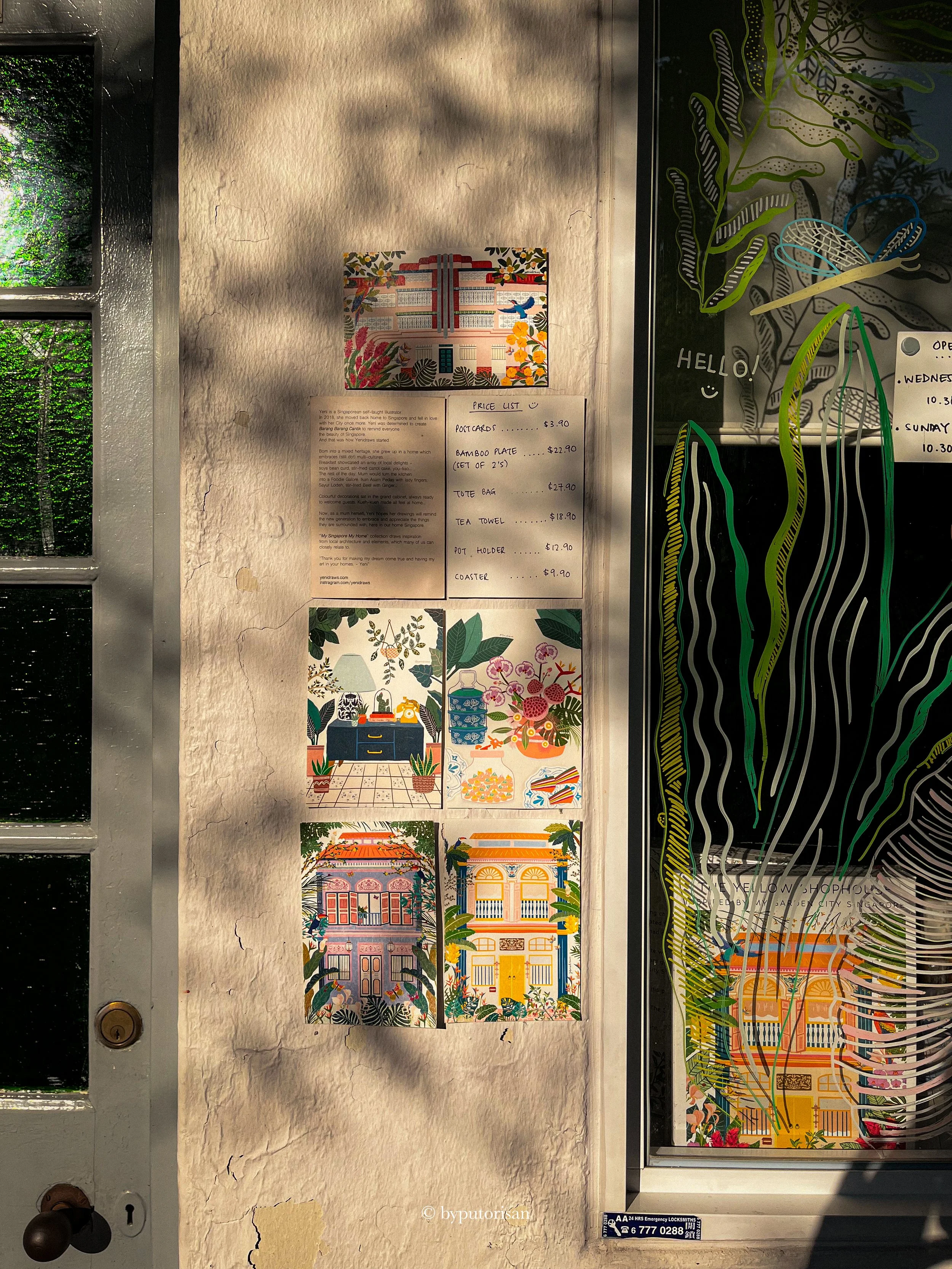 A display of colorful illustrated postcards and artwork on a wall, with a small black and white sign at the bottom right indicating emergency locksmith contact information. The postcards are arranged vertically along a textured, slightly cracked wall, and there is a window with a floral or leafy design on the right side.