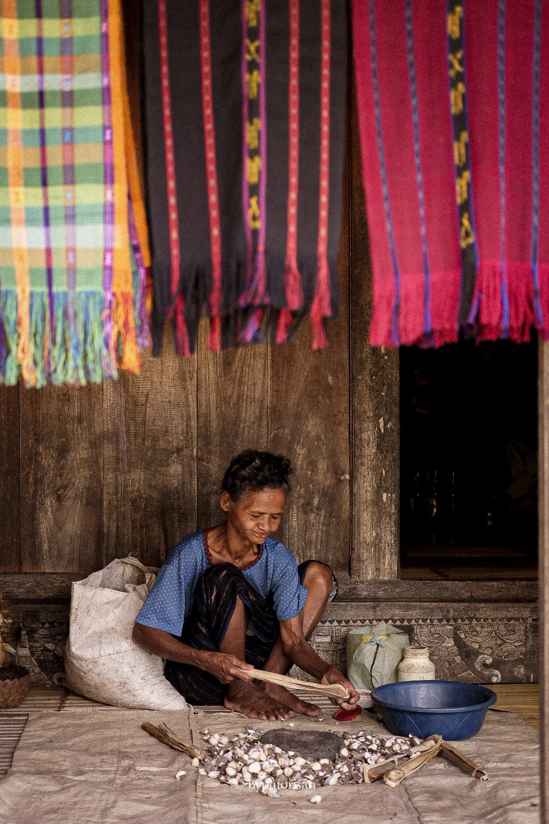 A woman sitting on the floor, peeling garlic, inside a wooden house with colorful fabrics hanging above her.