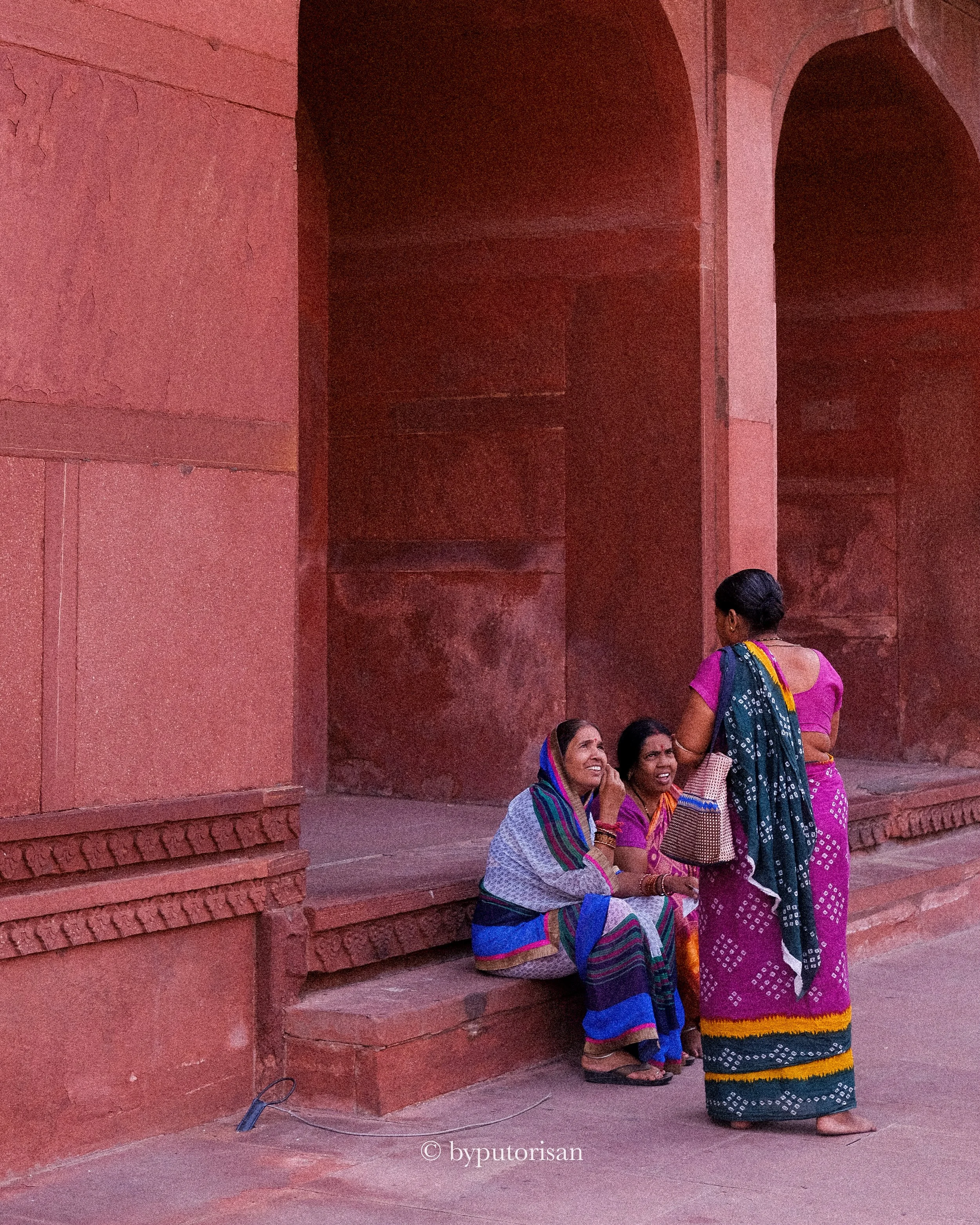 Four women dressed in traditional Indian sarees and clothing are gathered outside a red brick building, sitting and standing near the wall, engaged in conversation.
