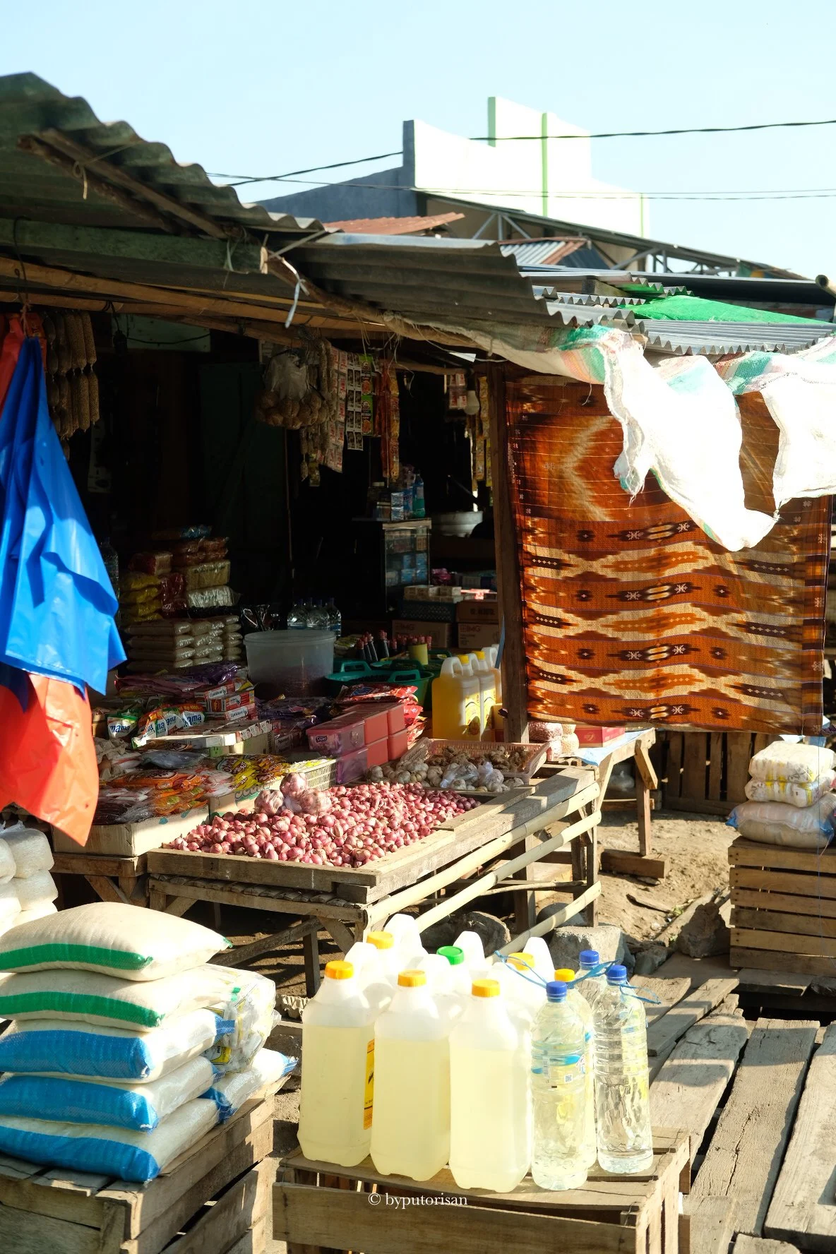 An outdoor market stall with various goods, including sacks of grains, bottles of liquids, bags of snacks, and a display of onions.