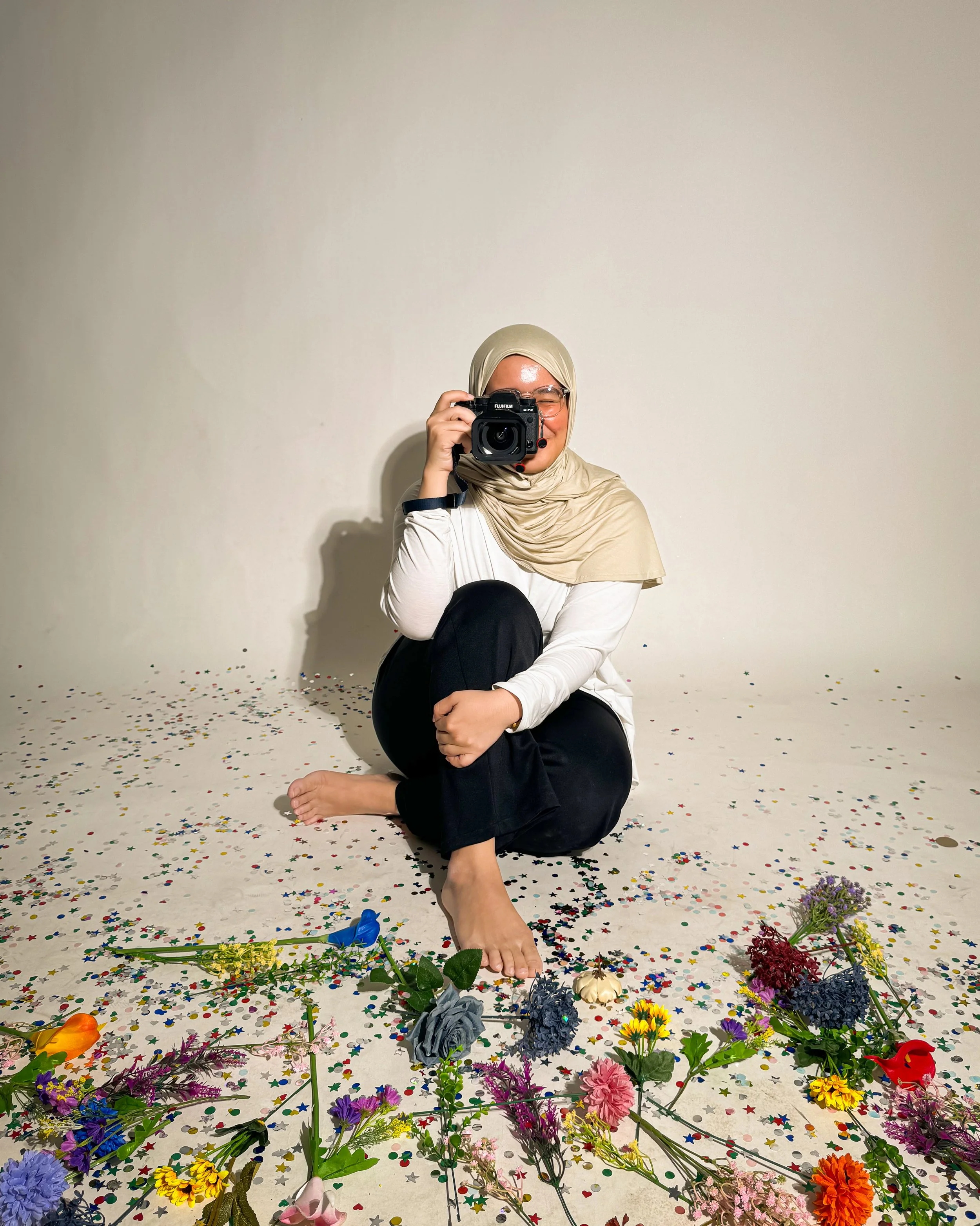 A woman taking a self-portrait with a camera, sitting on the floor surrounded by colorful flowers and confetti.