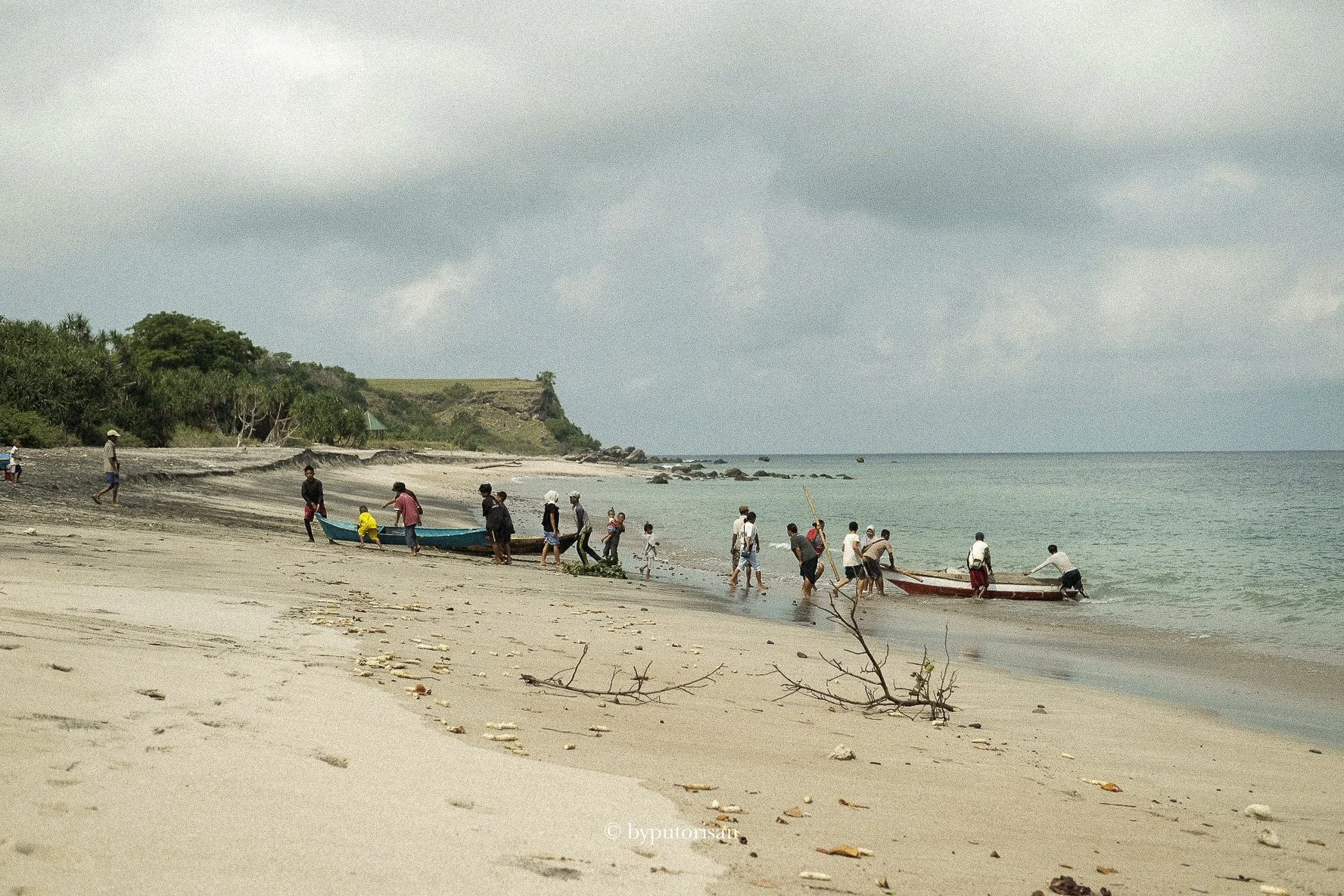 Group of people on a sandy beach launching small boats into the water under cloudy skies, with green hillside in the background.
