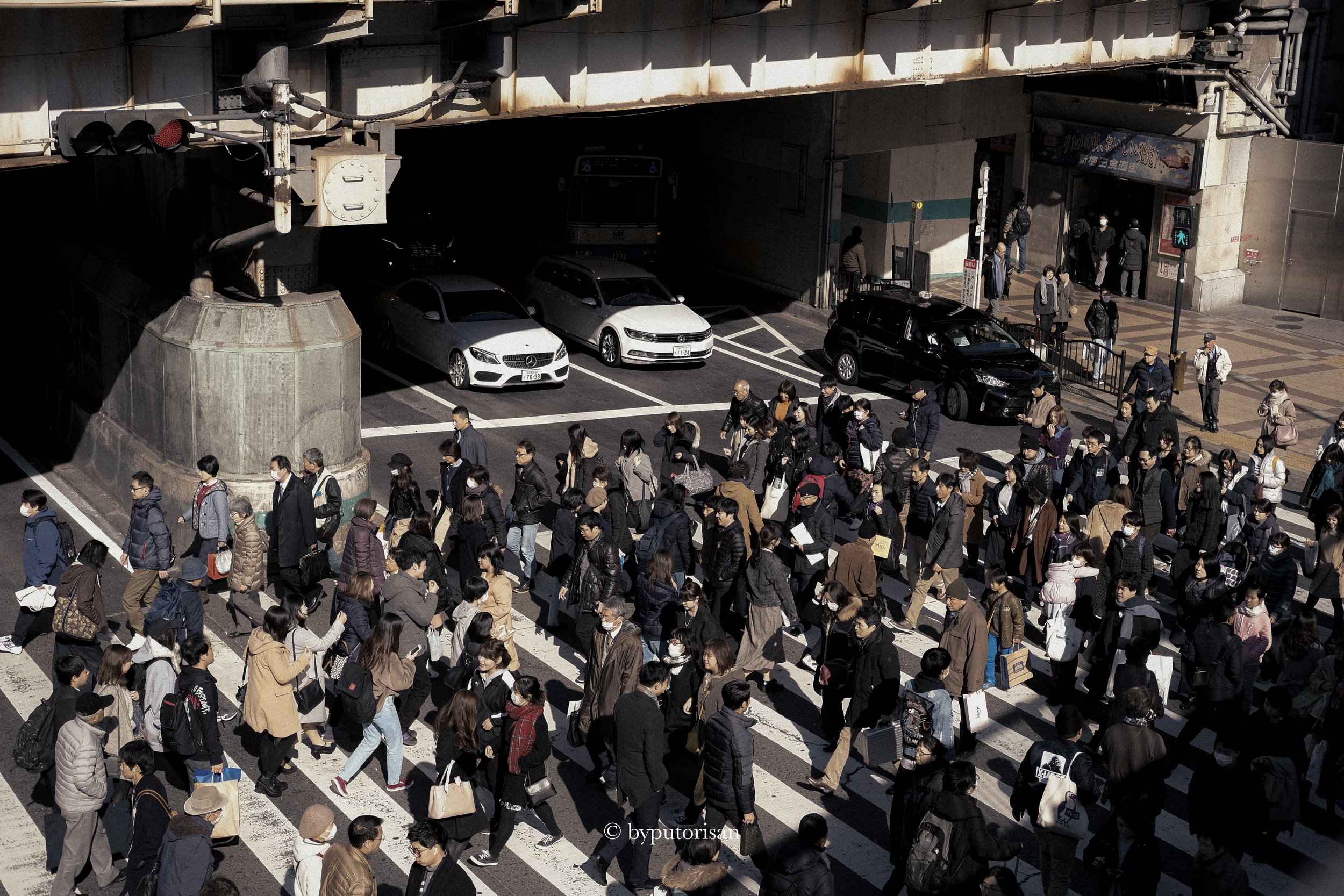 People crossing a busy city street at a crosswalk near an underpass with parked cars and a traffic signal.