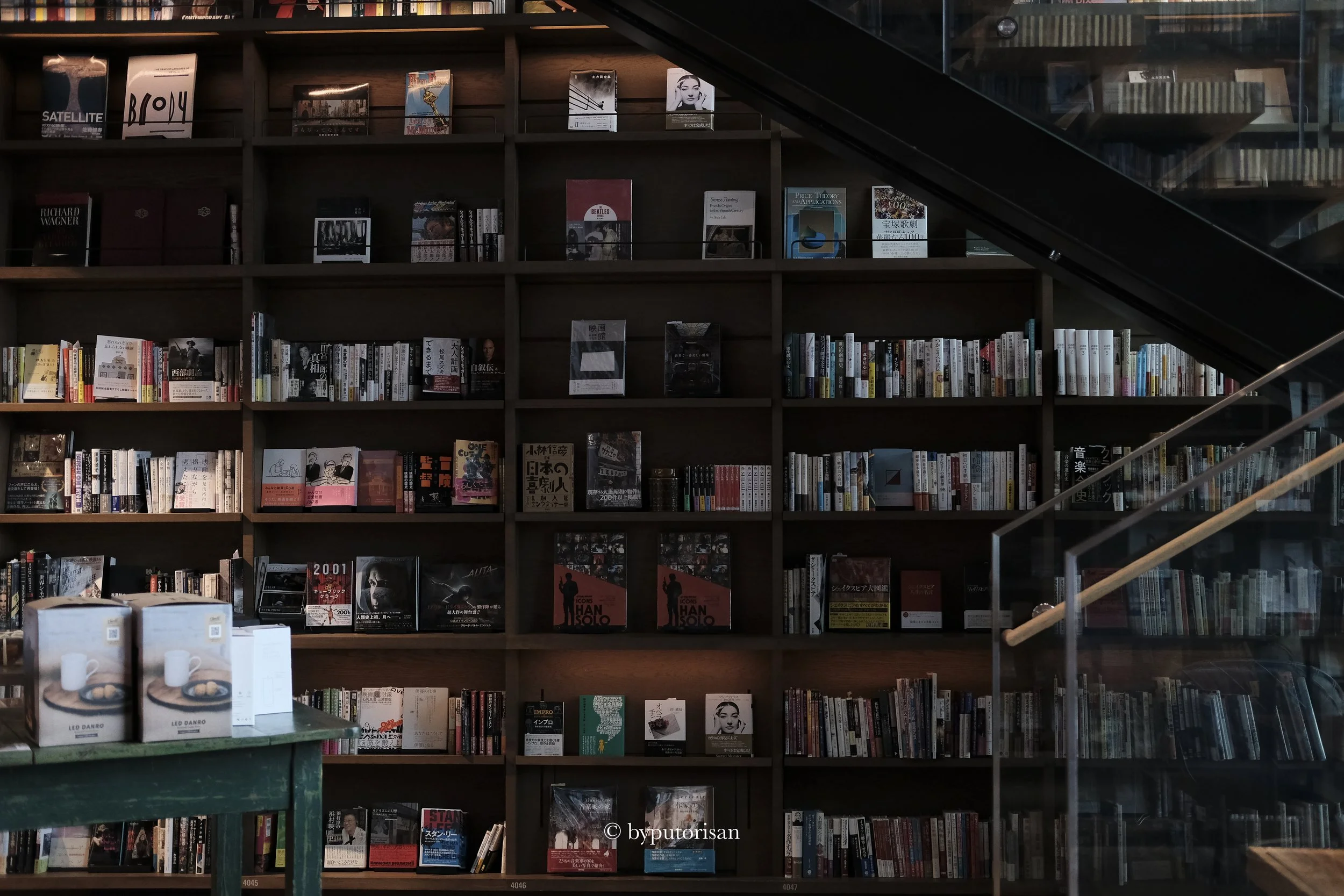 Interior view of a bookstore or library with wooden shelves filled with books. A staircase with glass railing is visible on the right side.