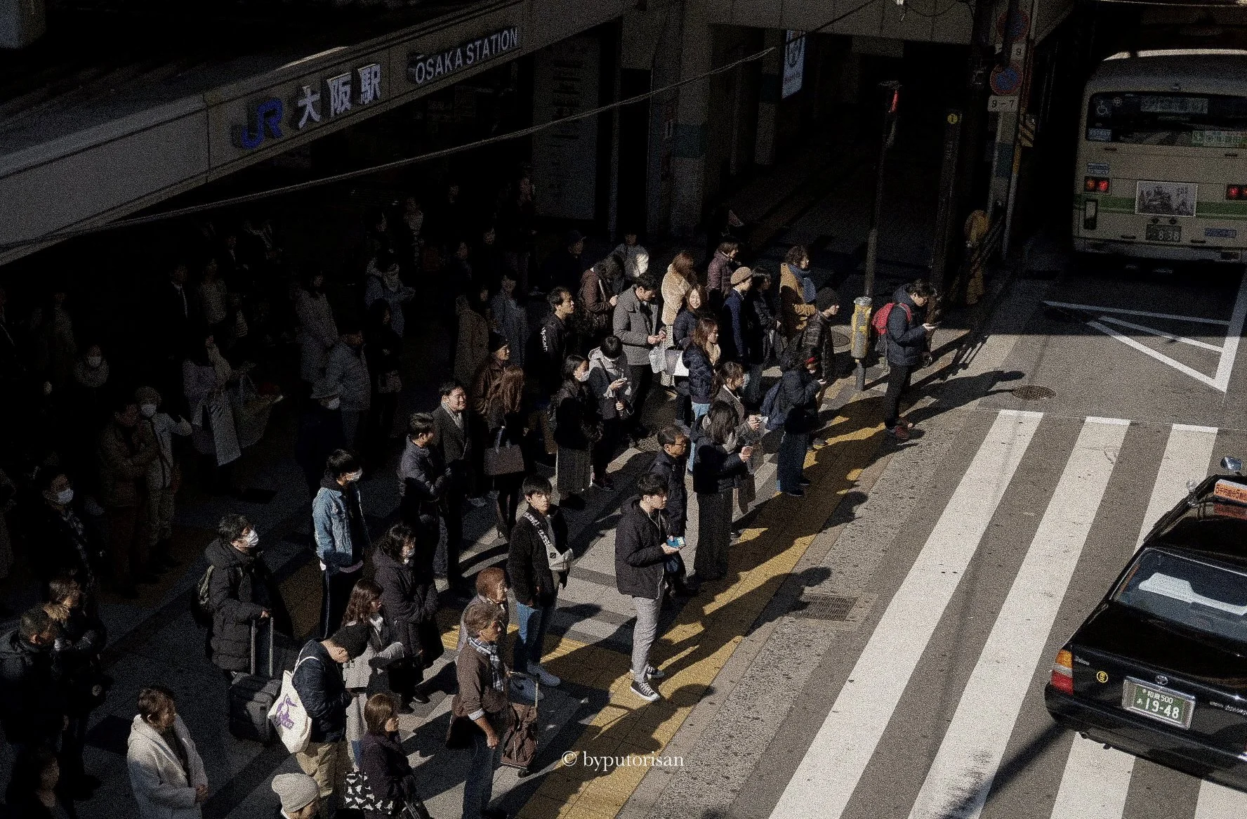 A long line of people waiting at a crosswalk outside Osaka Station in Japan, some wearing face masks, as a bus approaches in the background.