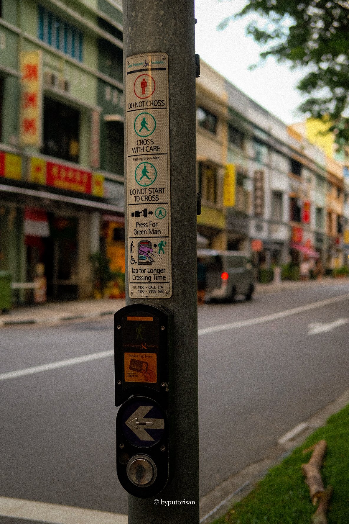 A pedestrian crossing control pole with instructions for crossing safety, including do not cross, cross with care, do not start to cross, press button for green man, and tap for longer crossing time, located on a city street with buildings and trees 