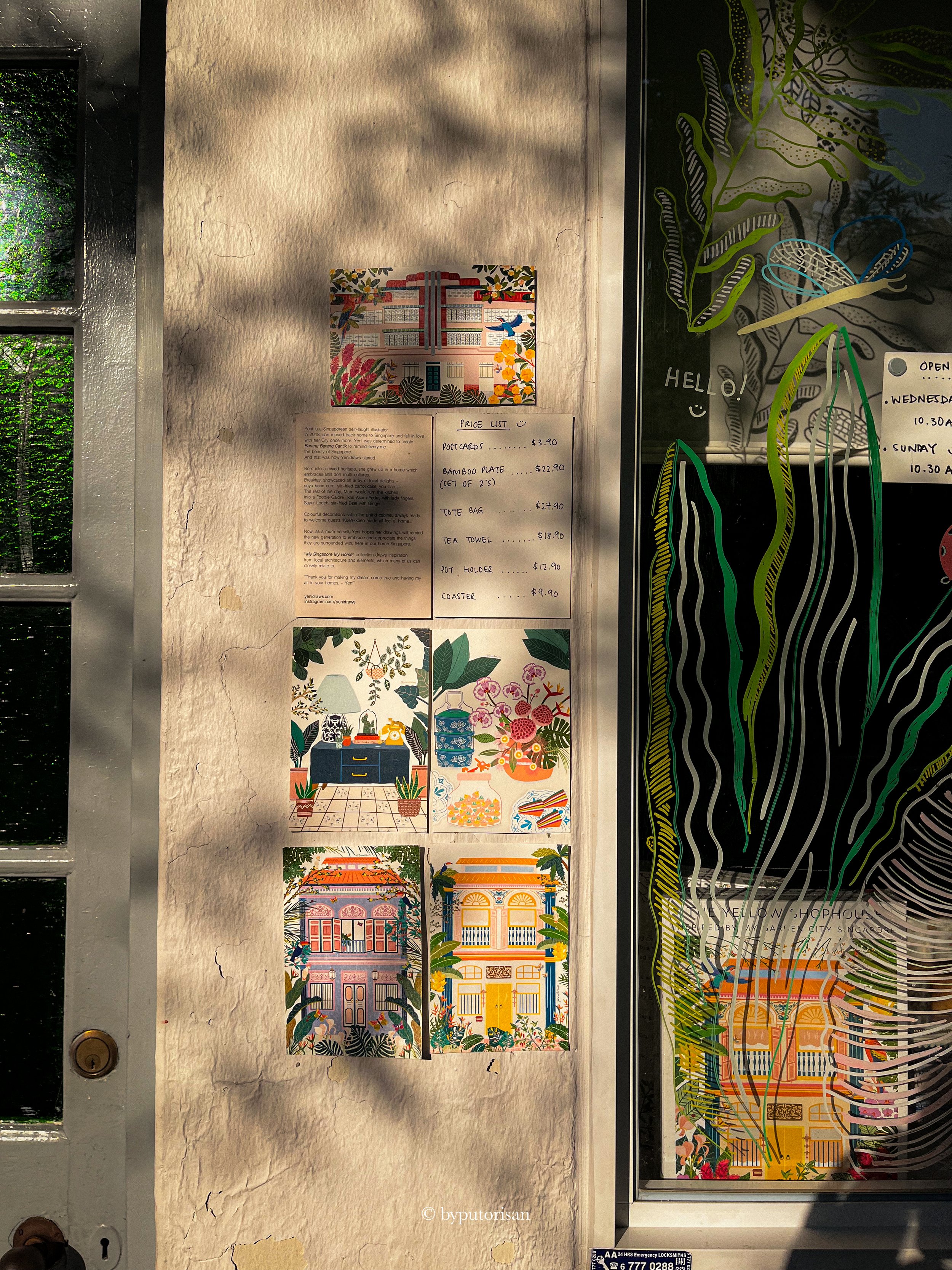 Decorative wall with artwork, a handwritten price list for souvenirs, and a window with colorful illustrations of buildings and plants.
