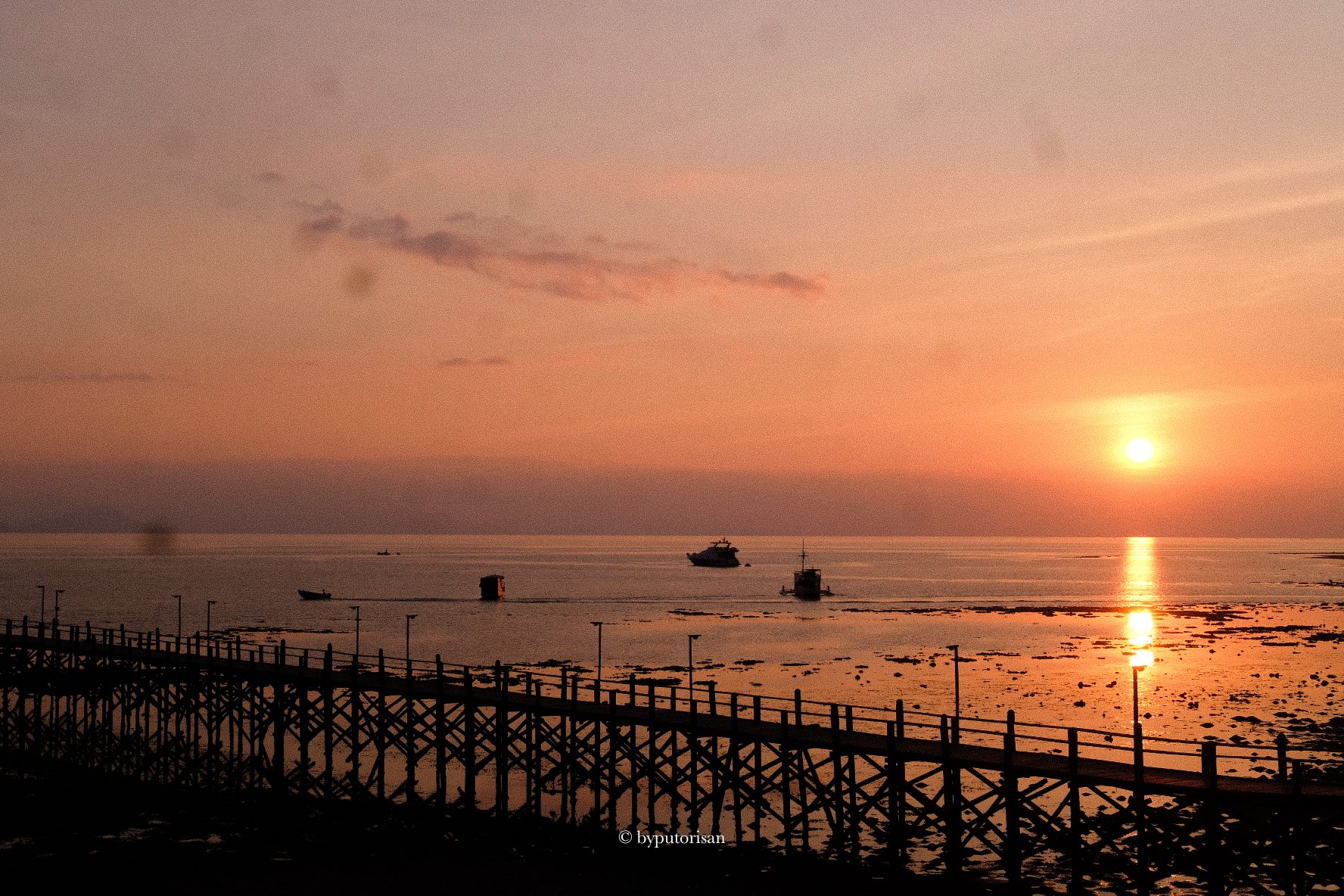 Sunset over the ocean with boats and a wooden pier in the foreground, orange and pink sky with clouds.